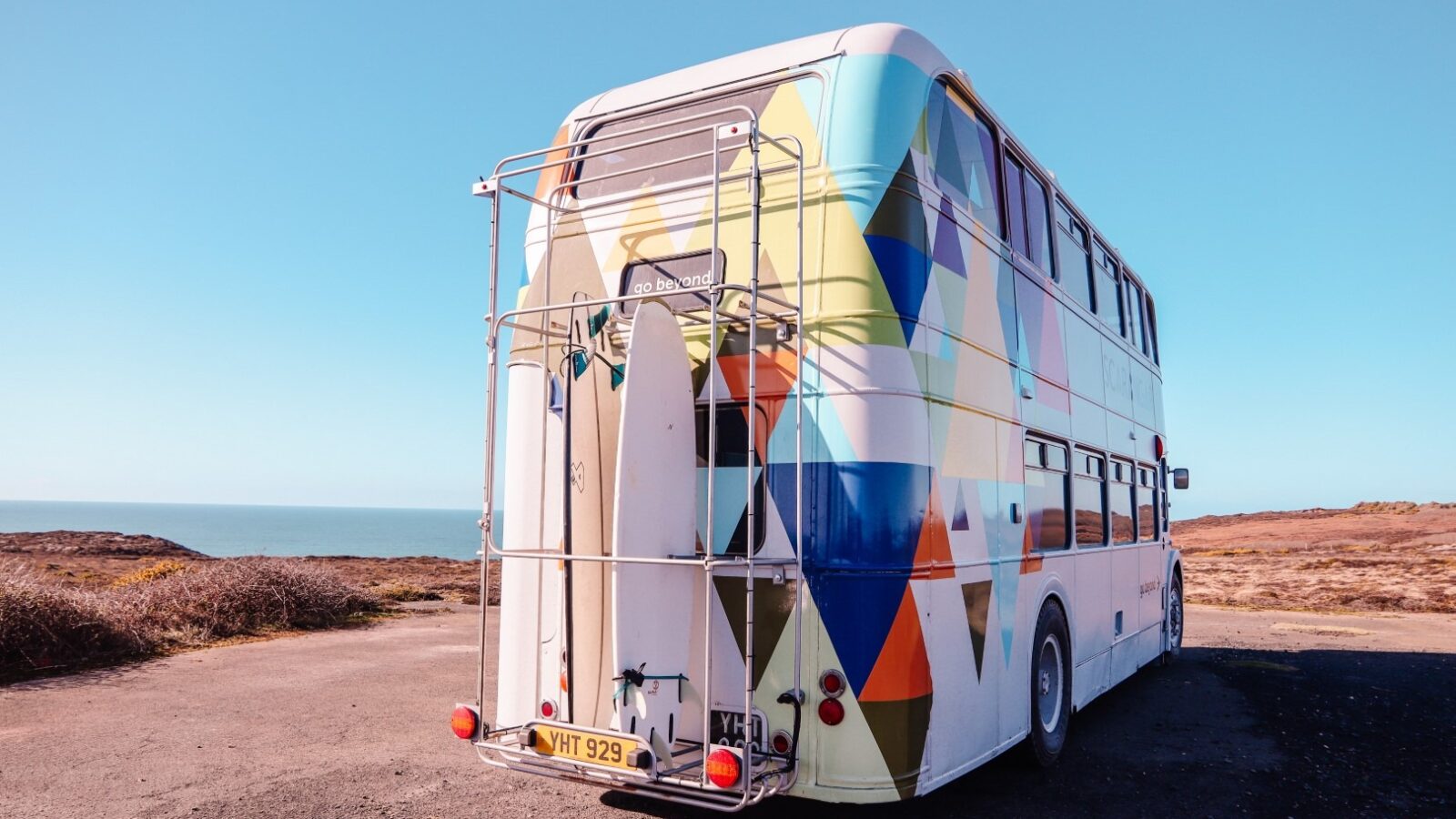 A colorful double-decker bus with geometric patterns, known as Scaragnar, is parked near a coastal area. Two surfboards are attached to a rack on the back of the bus. The sky is clear and blue, with the coastline visible in the background.