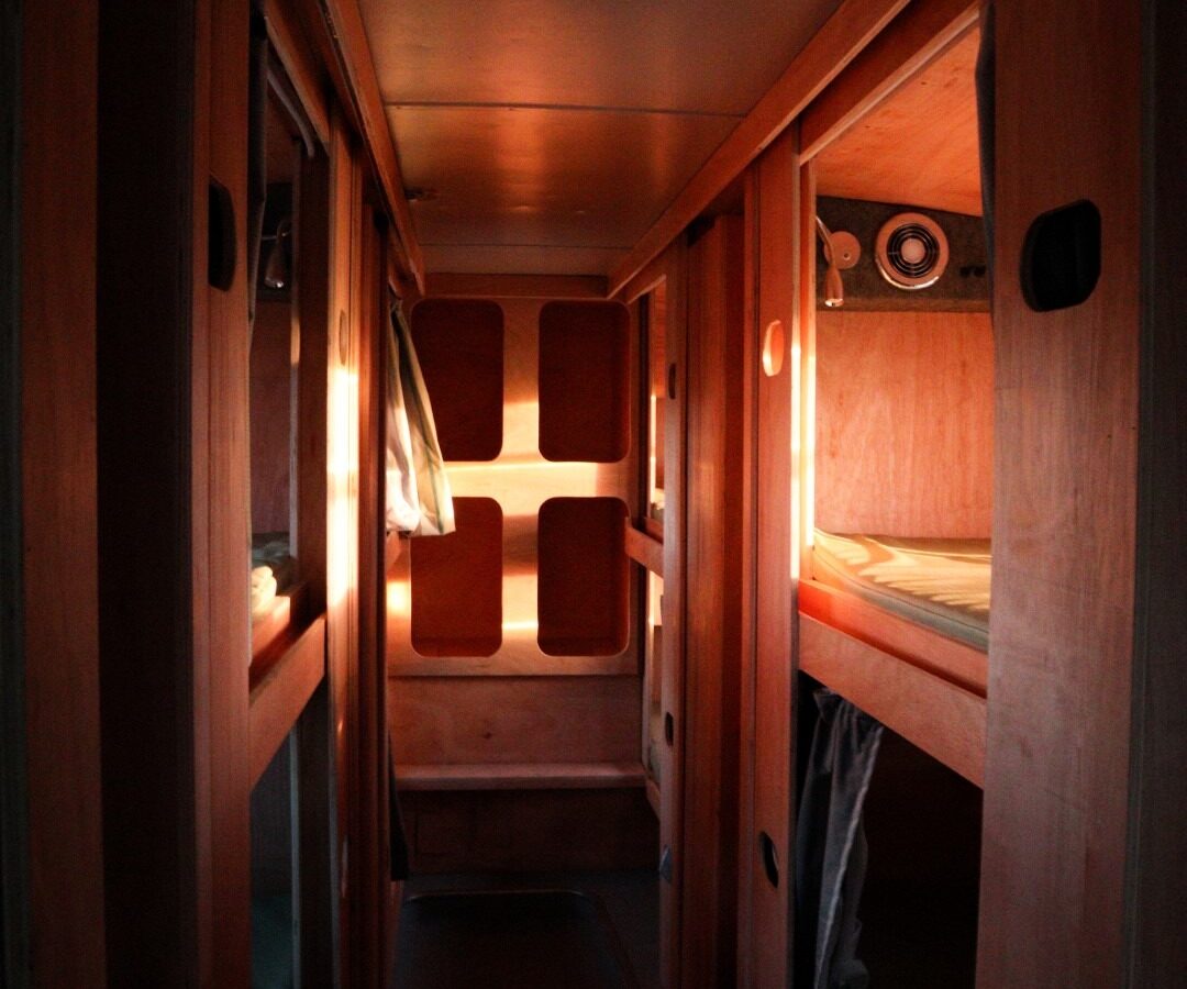 A narrow corridor inside a dimly-lit Scarangar cabin with wooden walls. Bunk beds with small windows are built into each side, and soft light filters in from the end of the corridor. The space looks cozy and compact, reminiscent of a sleeper train or hostel.