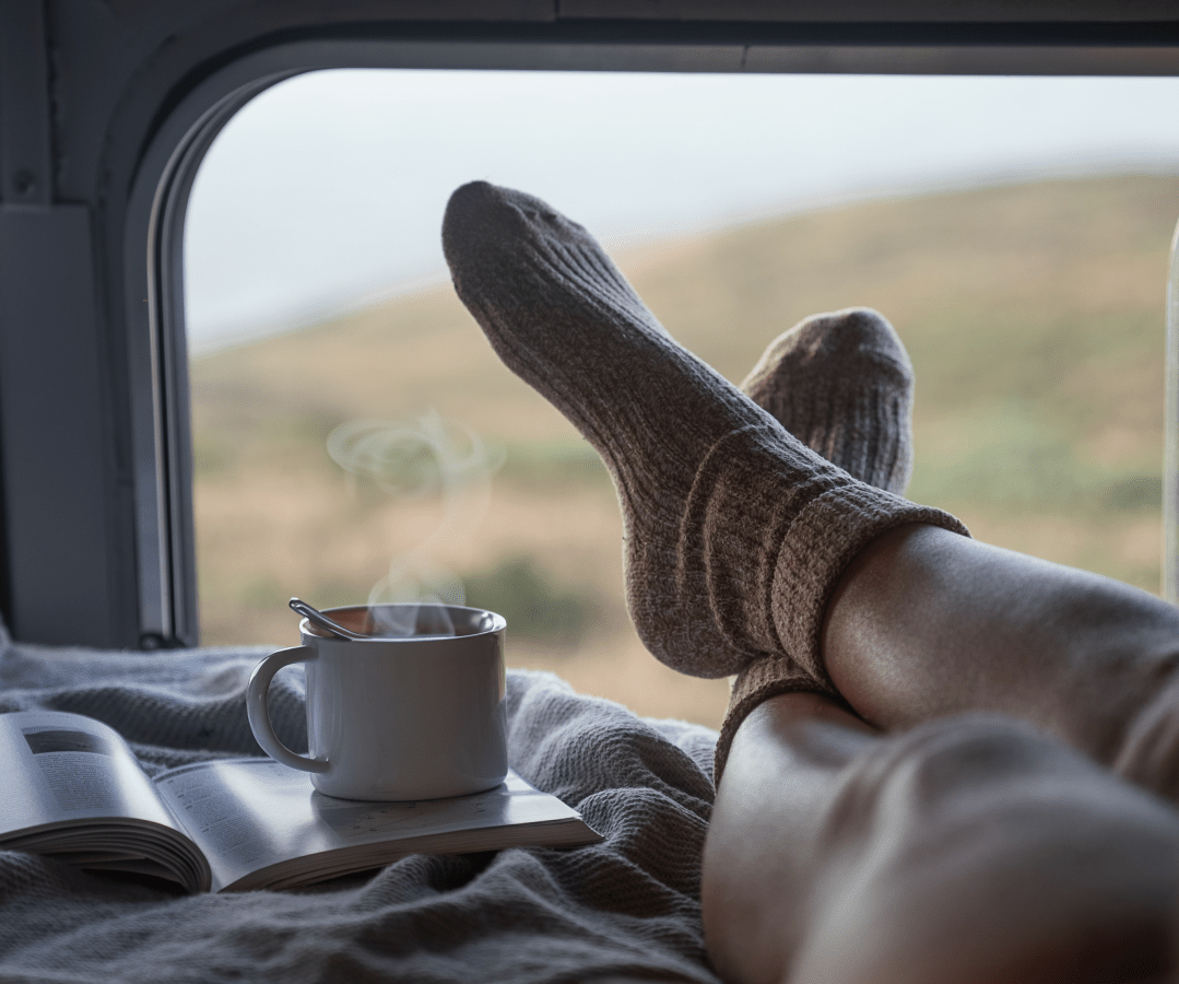 A person wearing cozy wool socks is relaxing with their feet up inside a Scarangar camper van, with a steaming mug of coffee or tea and an open notebook nearby. The view outside the window shows a blurred natural landscape.