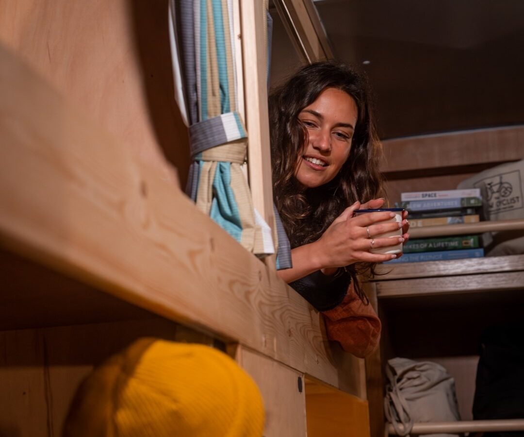 A woman with long curly hair, wearing an orange sweater, lies on the top bunk of a wooden Scaragnar bunk bed, holding a mug and smiling at a person in a yellow beanie on the lower bunk. Shelves with books and a backpack are visible in the background.