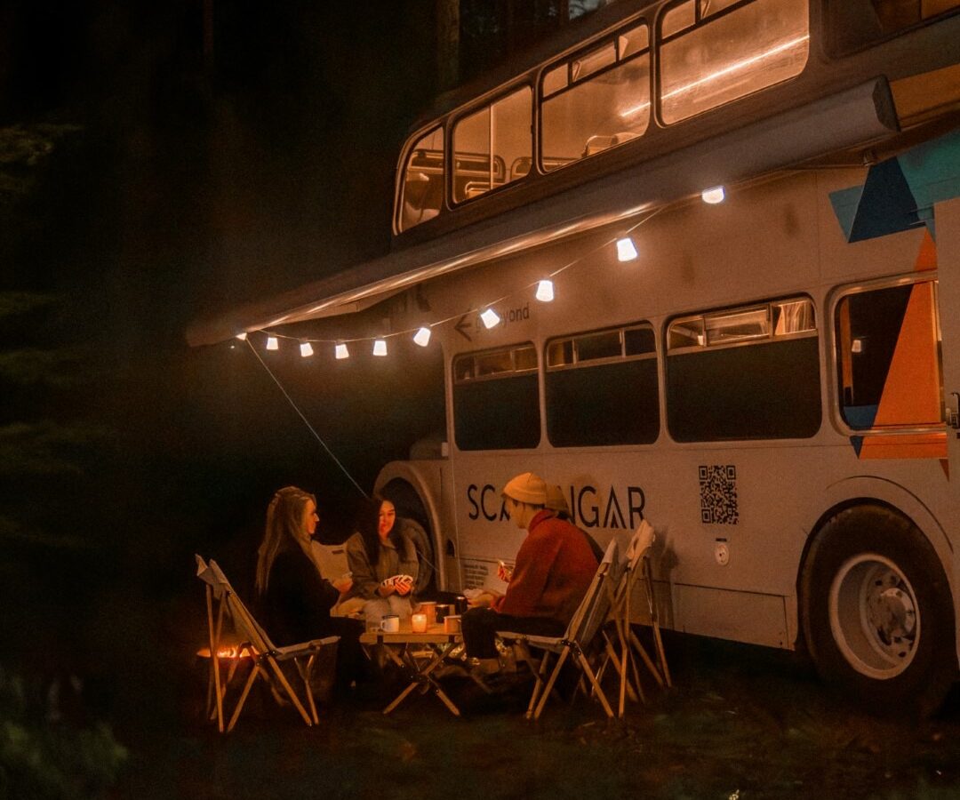 Three people sit around a table under the illuminated awning of a two-level camper van labeled 