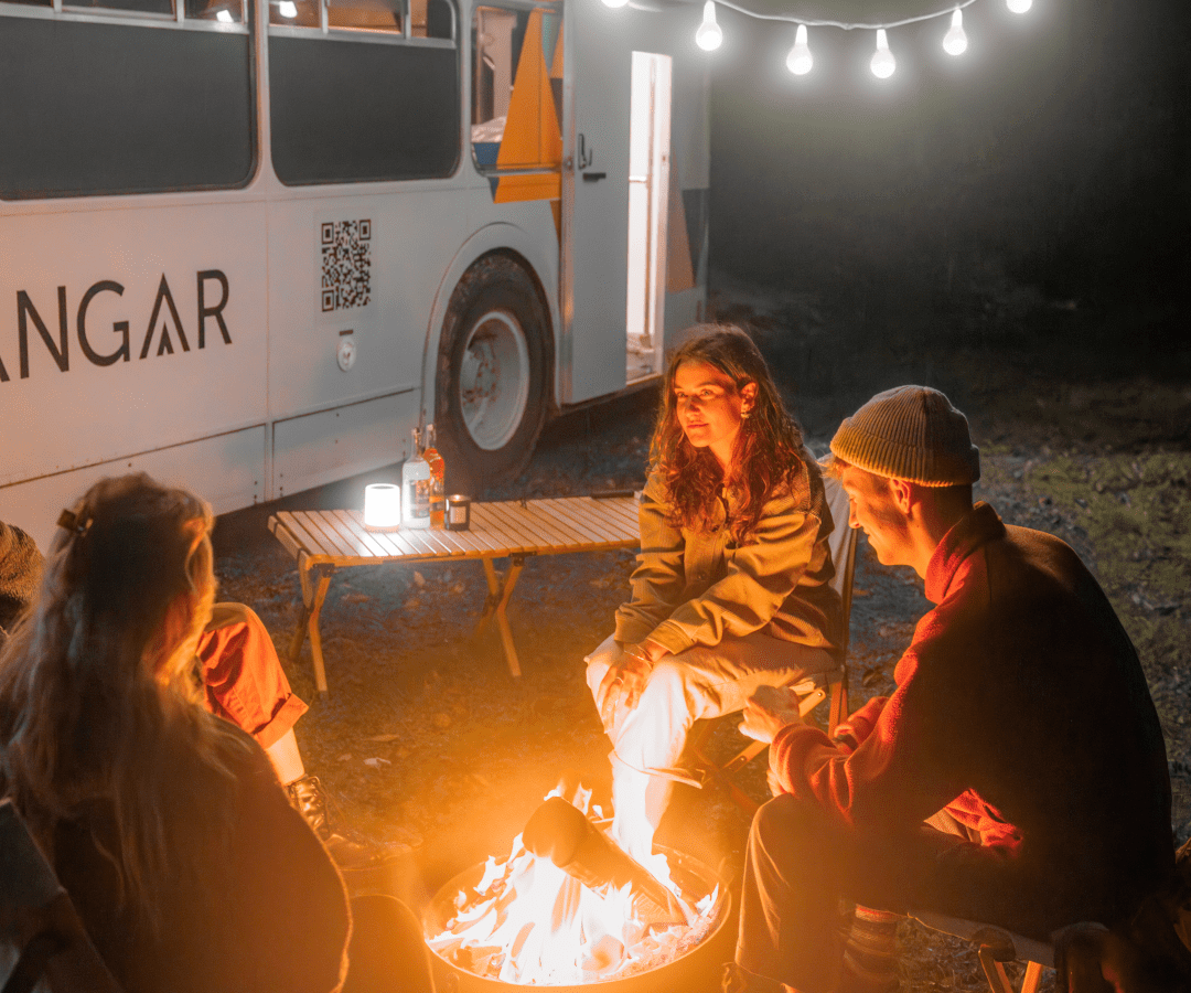 Three people sit around a campfire in the evening, outside a well-lit Scarangar camper van adorned with a string of lights overhead. Engaged in conversation and enjoying the warm fire, the scene is cozy, with a clear night sky visible in the background.