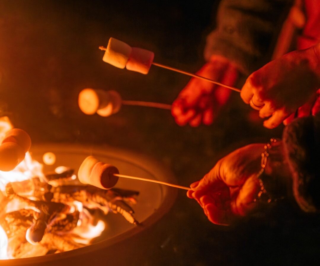 Hands holding sticks with marshmallows roast over an outdoor campfire at night. The fire illuminates the scene, casting a warm glow on the marshmallows and sticks. Several people, including Scarangar enthusiasts, are visible, engaged in the timeless process of toasting marshmallows.