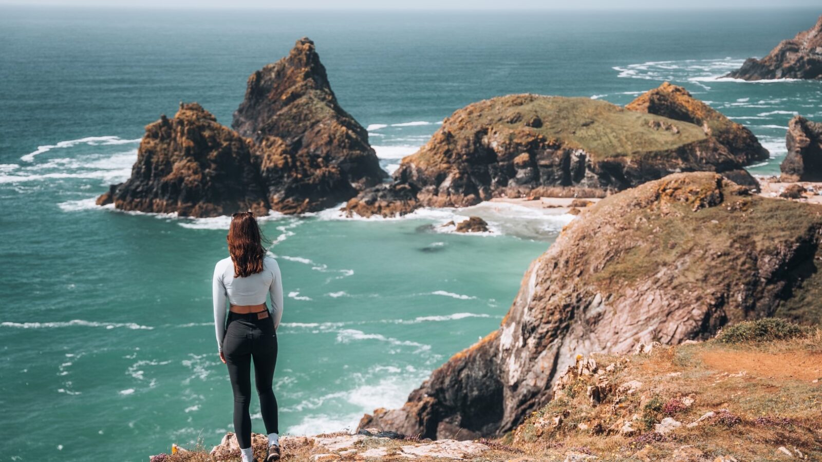 A woman stands on a rocky cliff in Scarangar, facing the sea, with her back to the camera. She is wearing a white top and black leggings. The view includes rugged coastal rocks and turquoise water, with waves gently crashing against the shore.