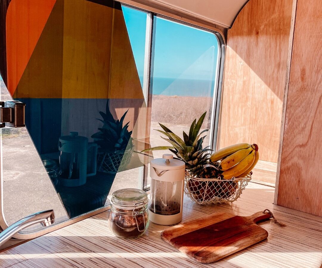 Interior view of a modern camper van featuring a stylish kitchen area. The countertop holds a French press, a jar, a wooden cutting board, and a basket filled with bananas and pineapples. The window offers a partial ocean view outside with colored glass panels, perfectly showcasing the Scarandar lifestyle.