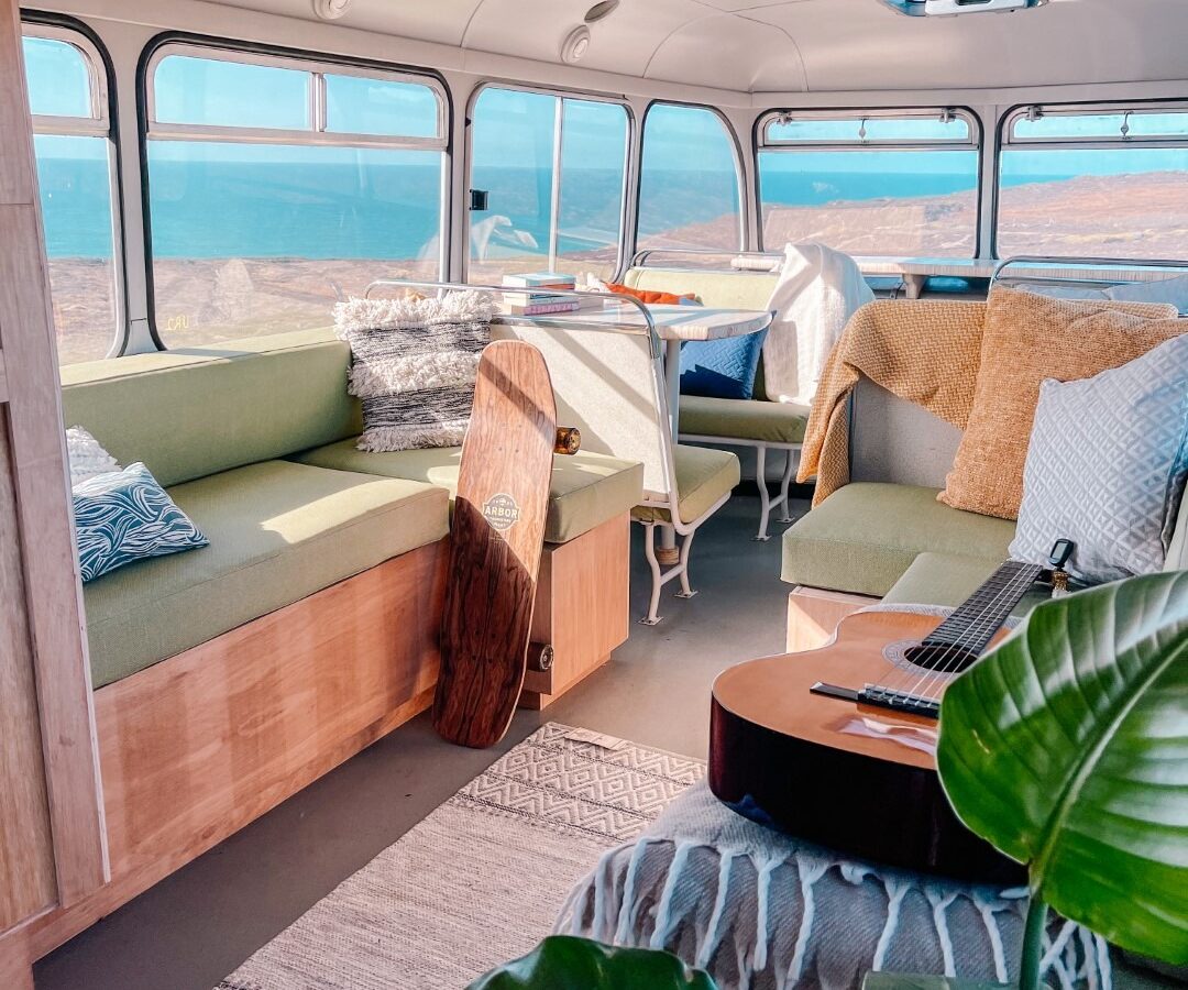 Interior of a converted Scarangar bus styled as a cozy living space with green benches, assorted pillows, and blankets. A skateboard leans against a bench, and an acoustic guitar rests on a cushion. Large windows reveal a scenic view of water and hills. A plant is in the foreground.