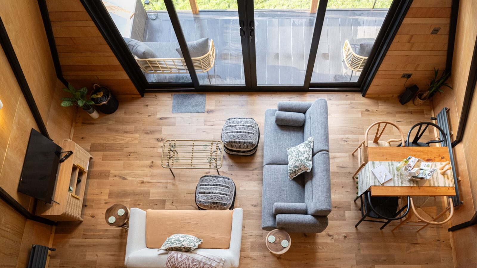 A cozy, modern living space as seen from above, reminiscent of the style at Sigrid Lodge, featuring a gray sofa, a white sofa, a glass coffee table, and two ottomans on a wooden floor. Adjacent is a dining area with a wooden table and chairs. Large glass doors open to a patio with chairs.