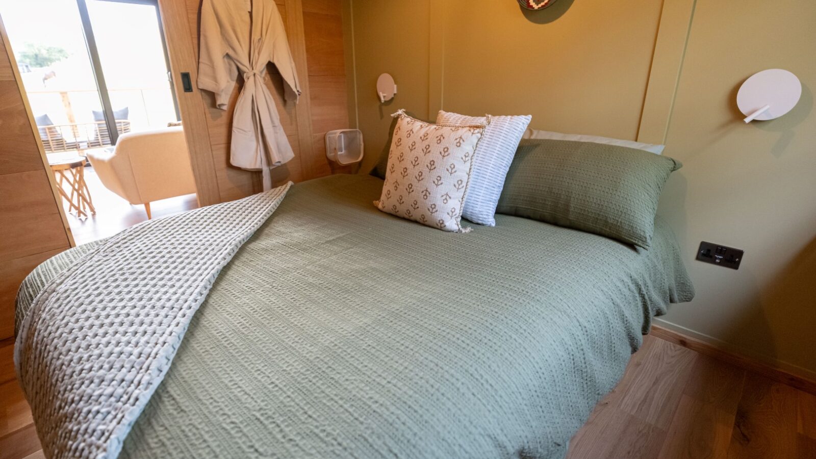 A cozy bedroom in Sigrid Lodge featuring a neatly made bed with green bedding and several decorative pillows. A white bathrobe hangs on the wooden wall, and natural light floods the room from a window in the background. The room has a serene and inviting atmosphere.