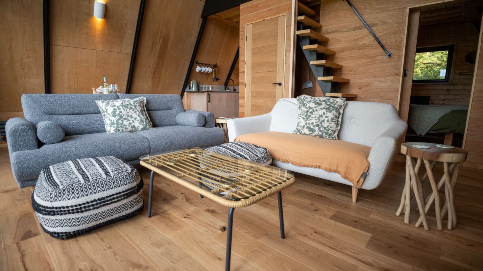 A cozy, modern living room with wooden walls and flooring reminiscent of a lodge. There are two stylish sofas with patterned pillows, a rattan coffee table, and a couple of poufs. A small side table and a staircase leading to an upper level are also visible. A bedroom is seen in the background.