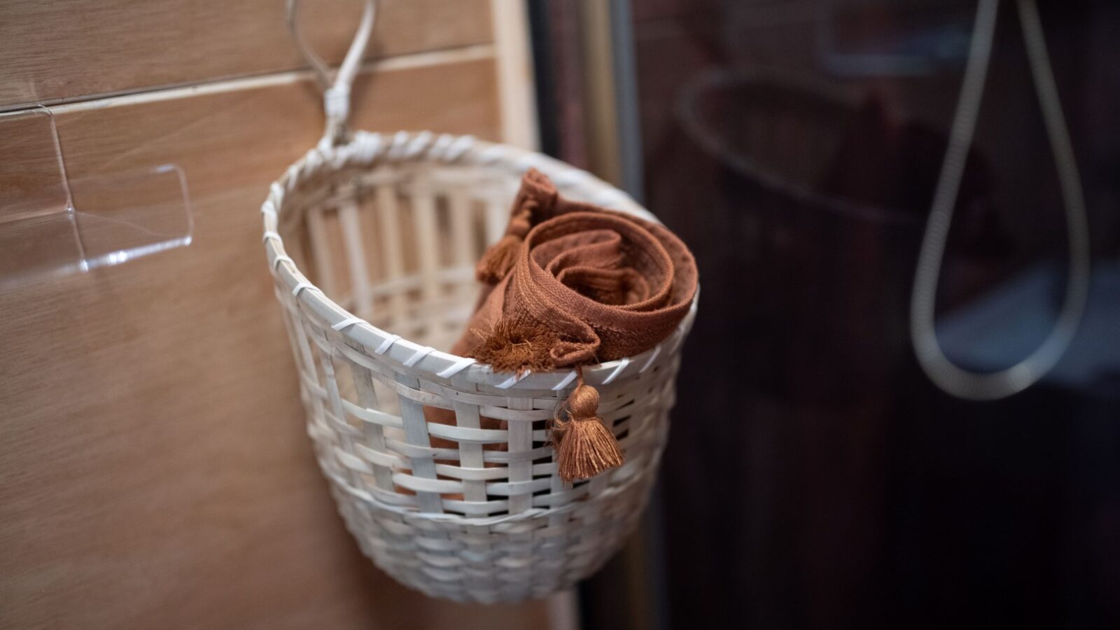 A small woven basket is hanging on a bathroom wall, containing neatly rolled brown towels with tassels, adding a touch of Sigrid Lodge charm. The basket has a loop for hanging, and the wall behind is tiled in light-colored tiles. A glass shower enclosure is partially visible in the background.