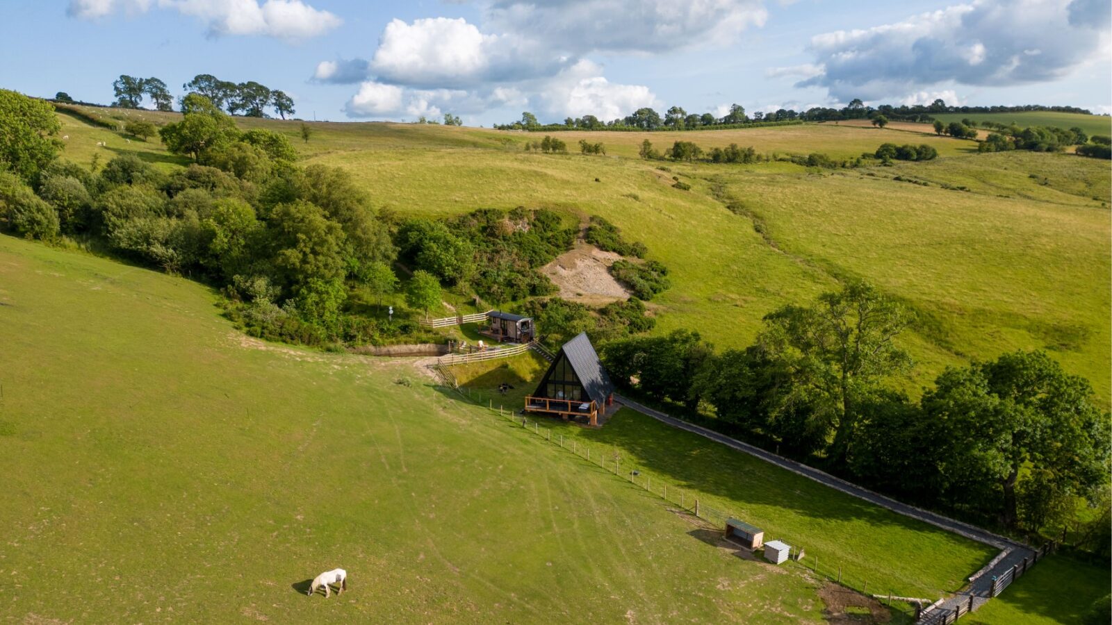 A rustic, triangular-shaped cabin known as Sigrid Lodge is nestled on a lush, green hillside surrounded by trees and fields. A lone white horse grazes in the foreground. Rolling hills and a partly cloudy sky create a serene, bucolic atmosphere.