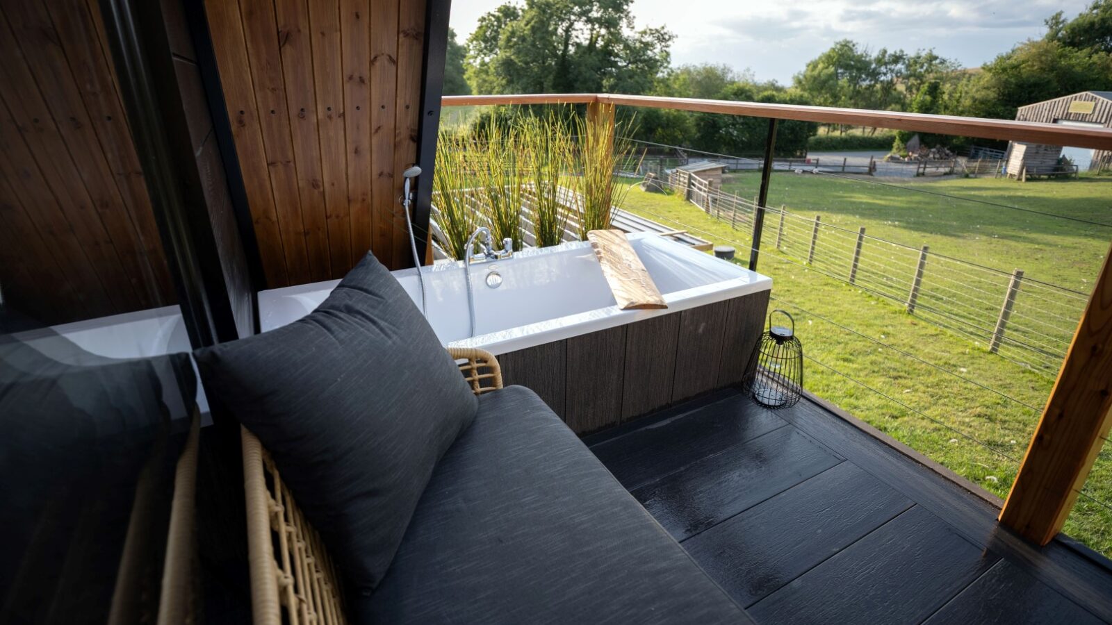 A cozy outdoor space at Sigrid Lodge with a modern patio sofa featuring dark cushions, a freestanding bathtub with a wooden plank for holding items, and a view of a lush green lawn surrounded by trees and a fence. The area is partially enclosed by wooden walls and glass panels.