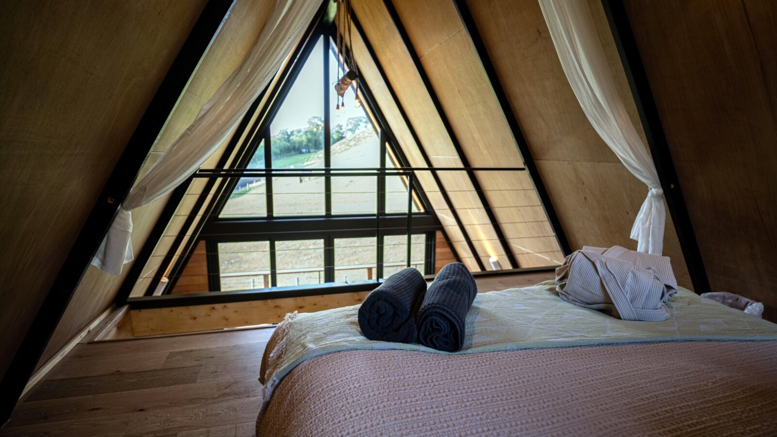 A cozy, A-frame attic bedroom in Sigrid Lodge features a large triangular window offering a scenic countryside view. The bed is neatly made with folded towels and a robe at the foot. Wooden floors and light curtains frame the window, creating a warm, inviting atmosphere.