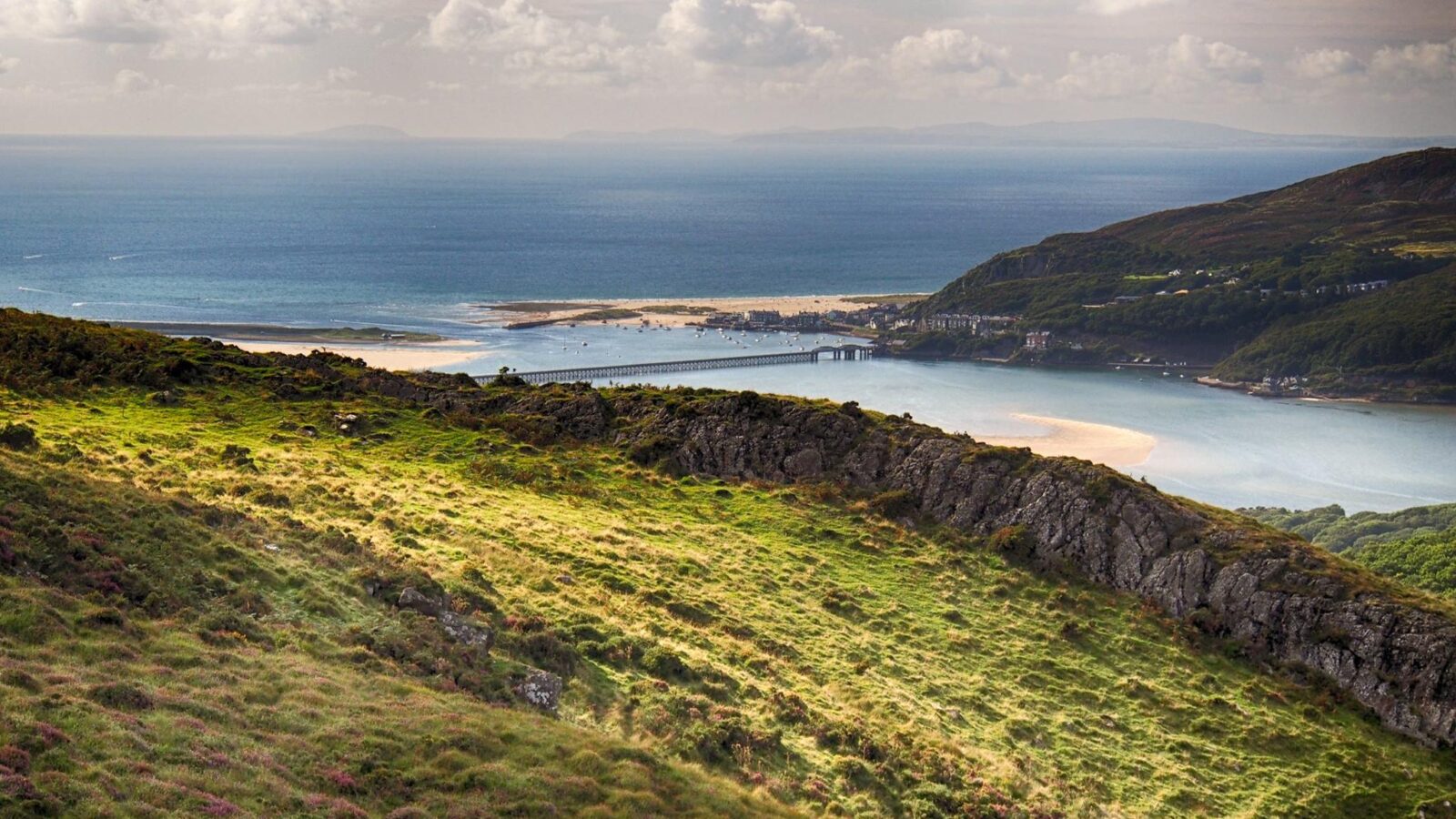 A scenic coastal landscape featuring rolling green hills in the foreground leads down to a distant bay. The bay includes a small town and a bridge crossing the water, offering a perfect hideaway. The sky is partly cloudy, and the blue sea extends to the horizon, reminiscent of Snowdonia's serene charm.