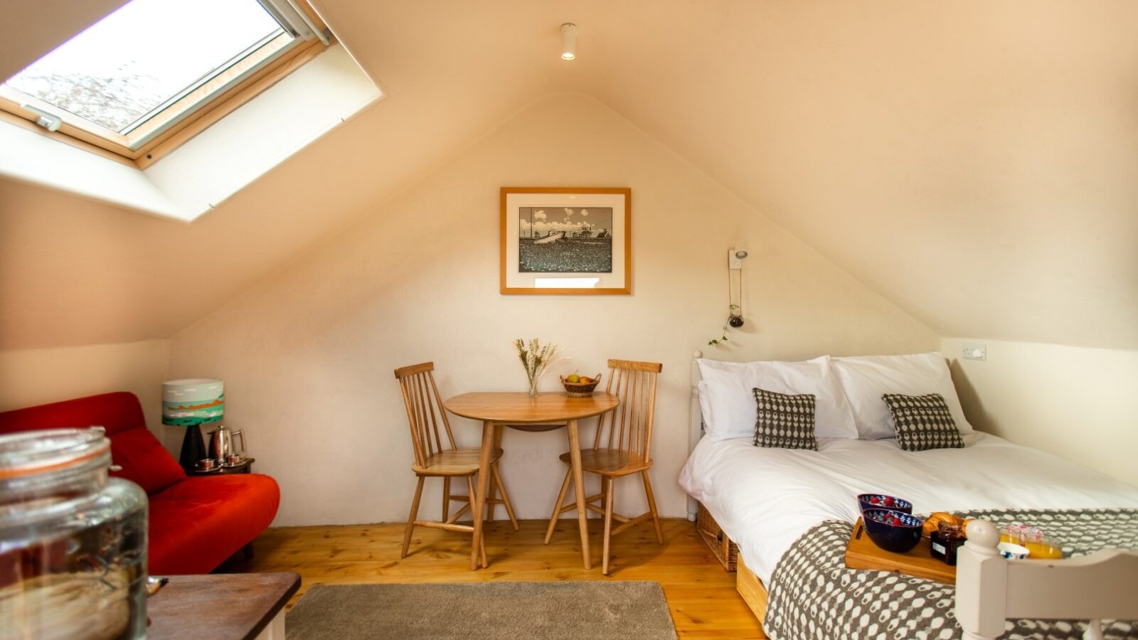 A cozy attic hideaway in Snowdonia features a skylight, a double bed with patterned pillows and a blanket, a small round wooden table with two chairs, a red sofa, and various decorations on a wooden floor. The white walls create a bright and airy atmosphere.