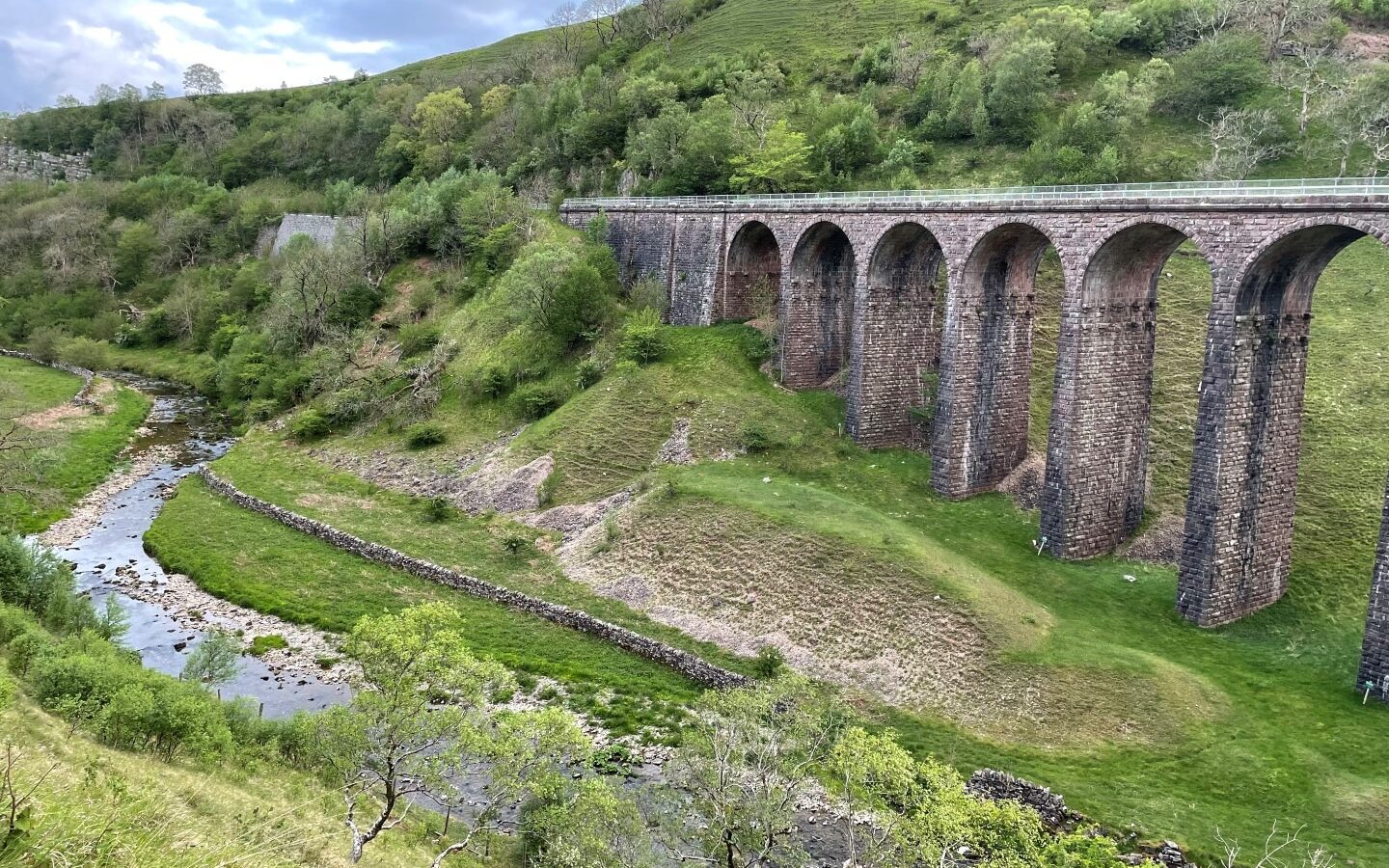 A scenic landscape featuring a stone arch bridge spanning a narrow, gently flowing river. The bridge stands tall with multiple arches, surrounded by lush green hills and a cozy holiday cottage. Patches of rocks and grassy slopes enhance the natural beauty while the sky is partly cloudy.