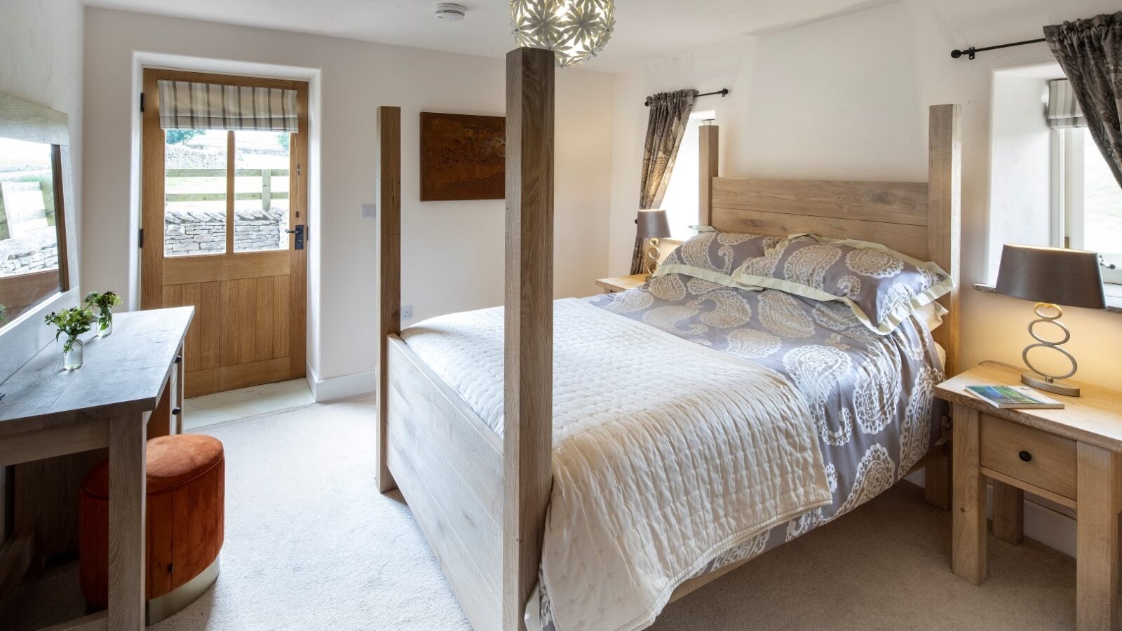 A cozy bedroom in a holiday cottage features a wooden four-poster bed adorned with patterned bedding. A wooden nightstand with a lamp is beside the bed. There is a small desk with a chair and plant. Two windows with dark curtains allow natural light in, while the door has a blind.