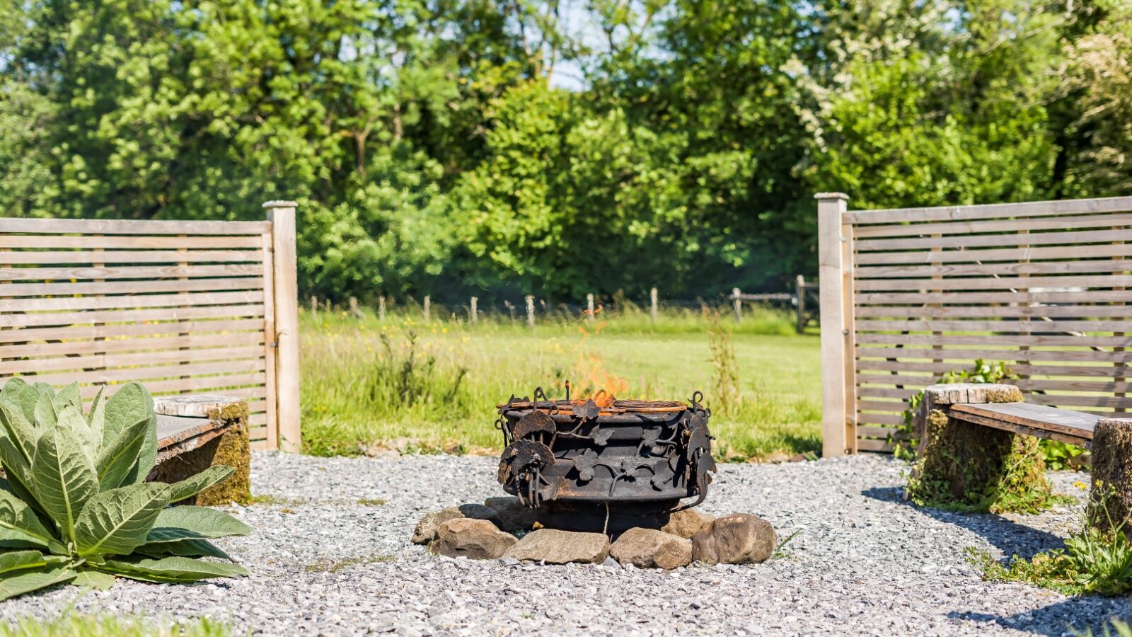 A cozy outdoor fire pit with flames, surrounded by stones, is the centerpiece of a gravel patio area at Erwain Escapes. Two rustic wooden benches are positioned on either side. The space is enclosed by wooden slatted fencing, with lush green trees and grass in the background, perfect for The Cabin ambiance.