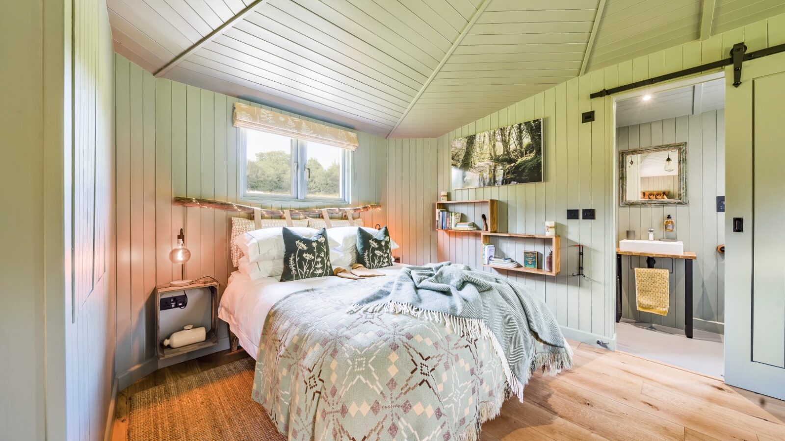 A cozy bedroom in The Cabin, with light wooden-paneled walls, features a bed adorned with patterned bedding and pillows. A window above the bed lets in natural light. There are wall shelves with decor, a bedside table with a lamp, and an adjacent bathroom with a sink and mirror by Erwain Escapes.