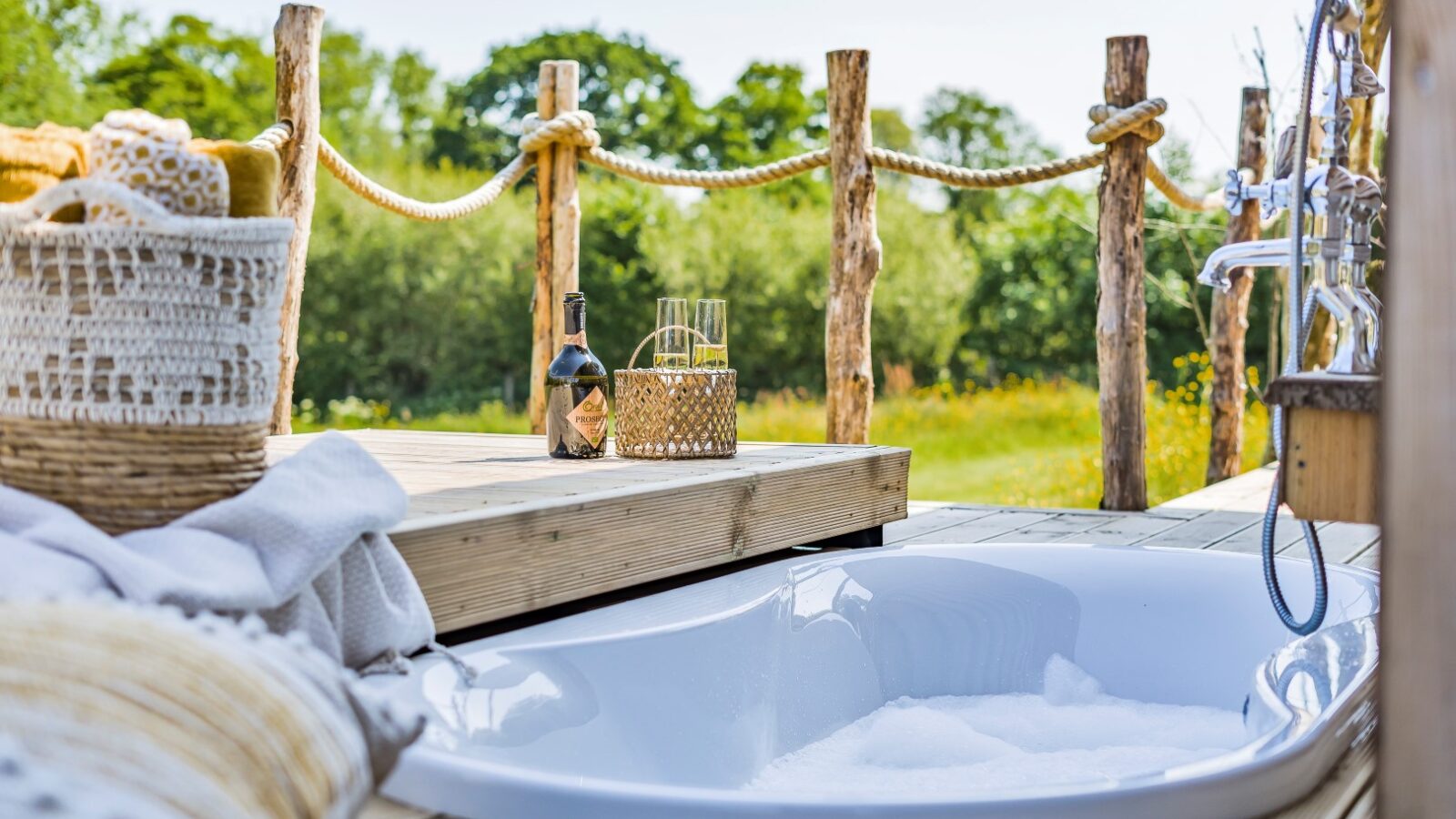 A sunken outdoor bathtub filled with bubbles is set on a wooden deck with a natural rope railing at Erwain Escapes. On the deck, there's a woven basket with towels, a bottle of wine, and a candle lantern. The background showcases lush green trees under a clear sky, embodying the perfect cabin retreat.