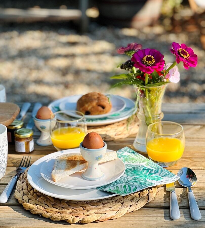 At the Green Hut, a breakfast table is set outdoors with sunlight. The table has a plate with a soft-boiled egg, toast, a glass of orange juice, cutlery, a small jar of jam, and a green napkin. Flowers in a vase brighten the setting as more food awaits in the background.