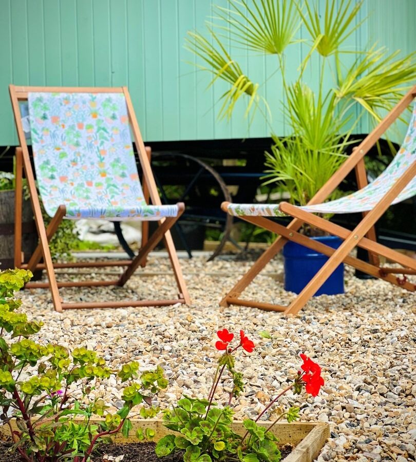 A small, tranquil garden area with two colorful deck chairs on a gravel surface. A green wooden wall forms the backdrop, reminiscent of the Green Hut's commitment to sustainable living, with a potted palm plant nearby. Red flowers bloom in a low, rectangular garden bed in the foreground.