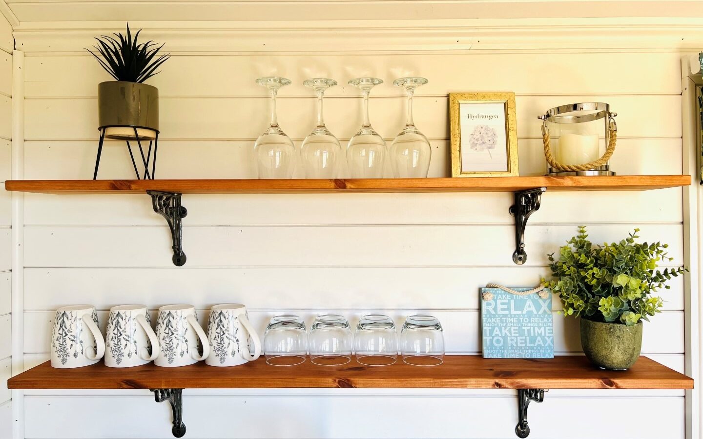 A cozy kitchen shelf setup with two wooden shelves against a white shiplap wall. The top shelf displays a plant, wine glasses, and a framed picture. The bottom shelf holds mugs and glasses. A sign reading 