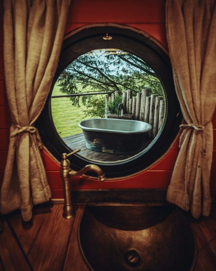 A round window framed by beige curtains looks out onto a patio at Fenny Castle, featuring a metal bathtub surrounded by a wooden fence. Inside, there is a bronze sink with a vintage-style faucet. Trees and the grassy expanse of The Orchard are visible in the background.