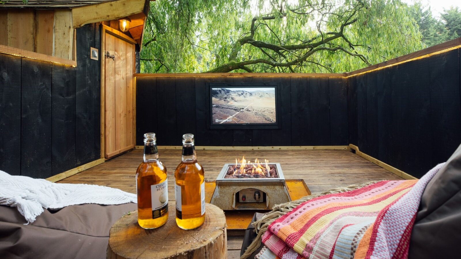 A cozy outdoor deck at Fenny Castle features a fire pit, a wooden side table with two bottles of beverage, cushioned seating with colorful blankets, and an outdoor TV mounted on the wall. Surrounded by greenery from The Orchard, the space exudes a rustic yet inviting atmosphere.