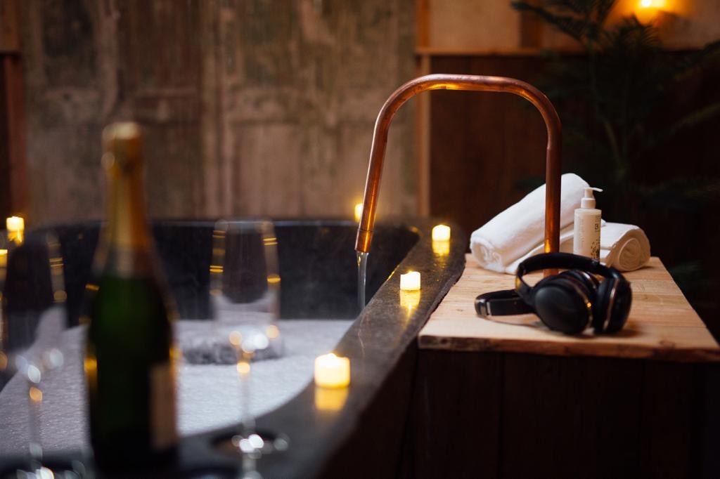 A luxurious wooden bathtub filled with bubbles and lit by numerous small candles is partially visible. On the rim, there are towels, a bottle of lotion, a pair of headphones, and nearby a bottle of champagne from Fenny Castle Orchard and two glasses, creating a cozy spa-like atmosphere.