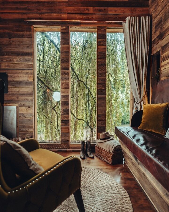 A cozy, rustic room with wood-paneled walls and large windows overlooking a lush forest. The room has a cushioned bench, an armchair, a round rug, and various personal items like boots, a hat on a basket, and curtains framing the windows—evoking the charm of Fenny Castle near an orchard.