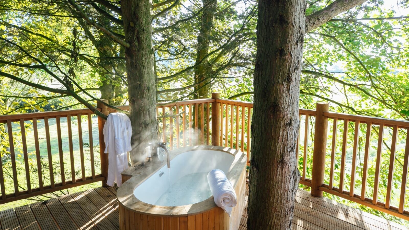 A serene outdoor wooden deck at Fenny Castle features a standalone bathtub filled with steaming water. A neatly rolled white towel rests on the tub's edge, and a white robe hangs on a railing. The area is surrounded by lush green trees, creating a tranquil, private setting.