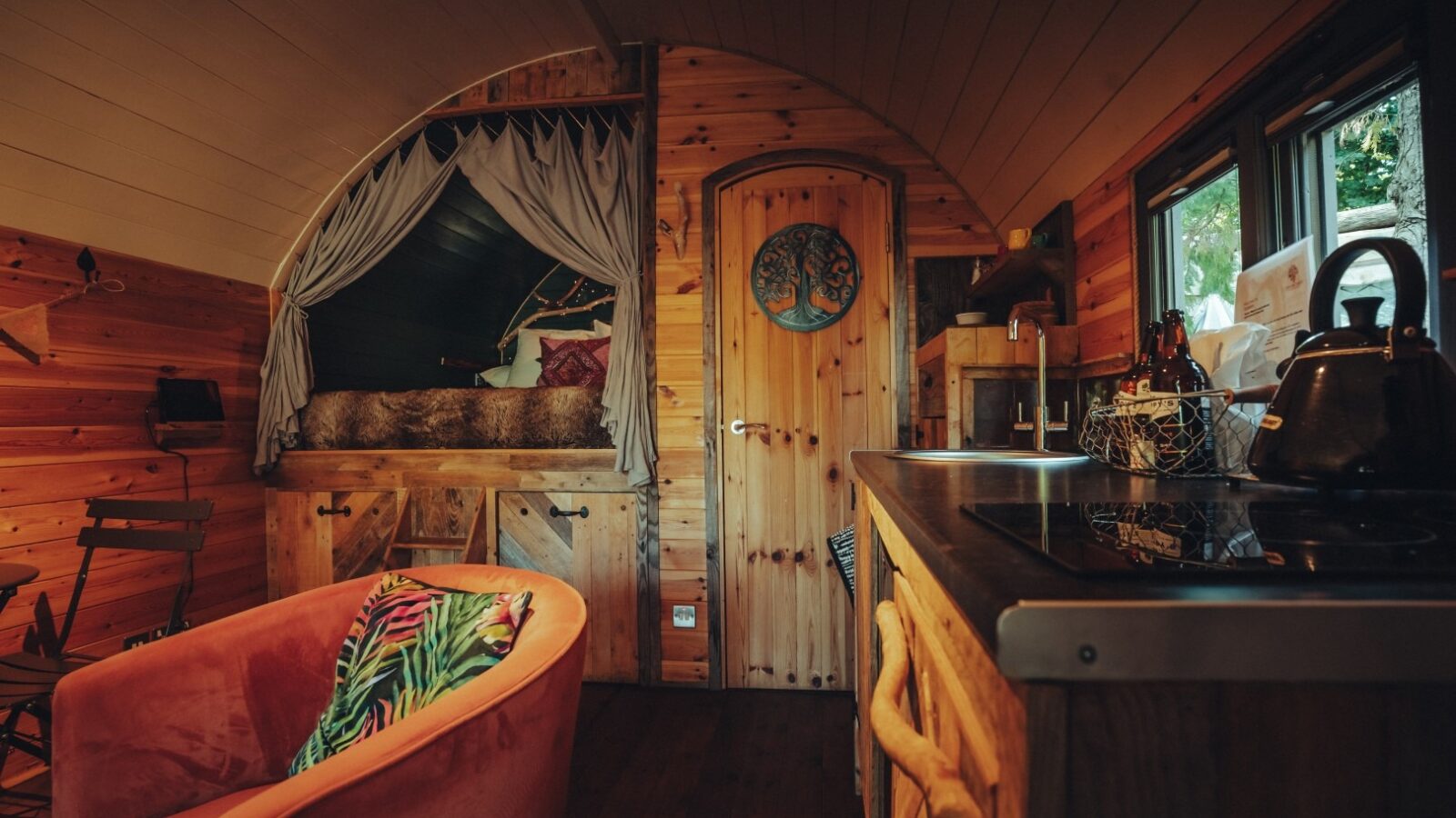 A cozy, rustic tiny house interior with wooden walls and ceiling, nestled near Fenny Castle. Features a kitchenette with a stove and kettle, a small table with two chairs, a couch adorned with colorful pillows, and a loft bed with curtains. Natural light streams in through a window overlooking The Orchard.