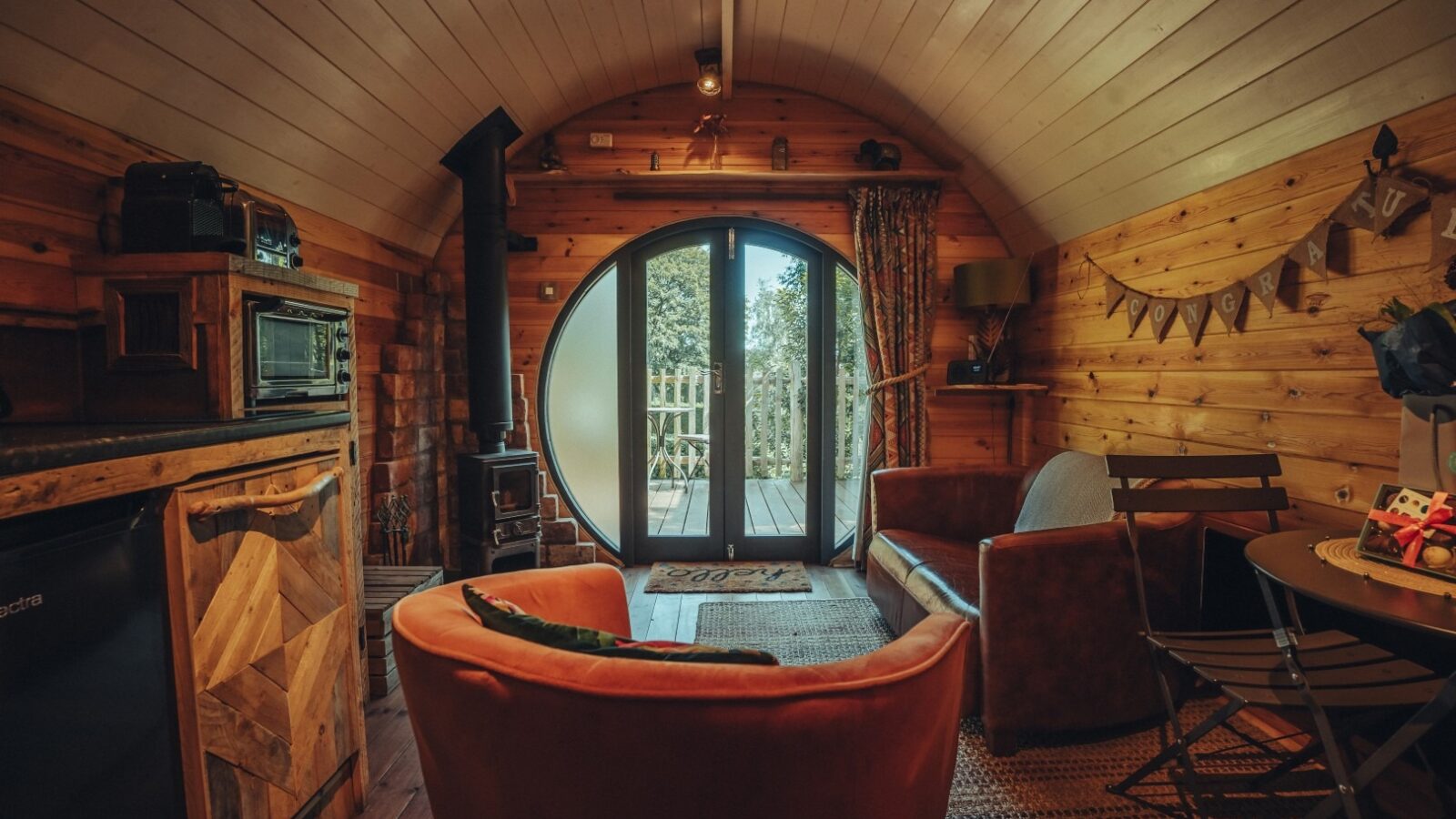 A cozy, oval-shaped wooden cabin interior featuring a comfy seating area with a brown leather sofa, orange armchair, and small table with chairs. Large arched glass doors lead to a deck outside. Fenny Castle's rustic decorations add warmth beside the wood-burning stove.