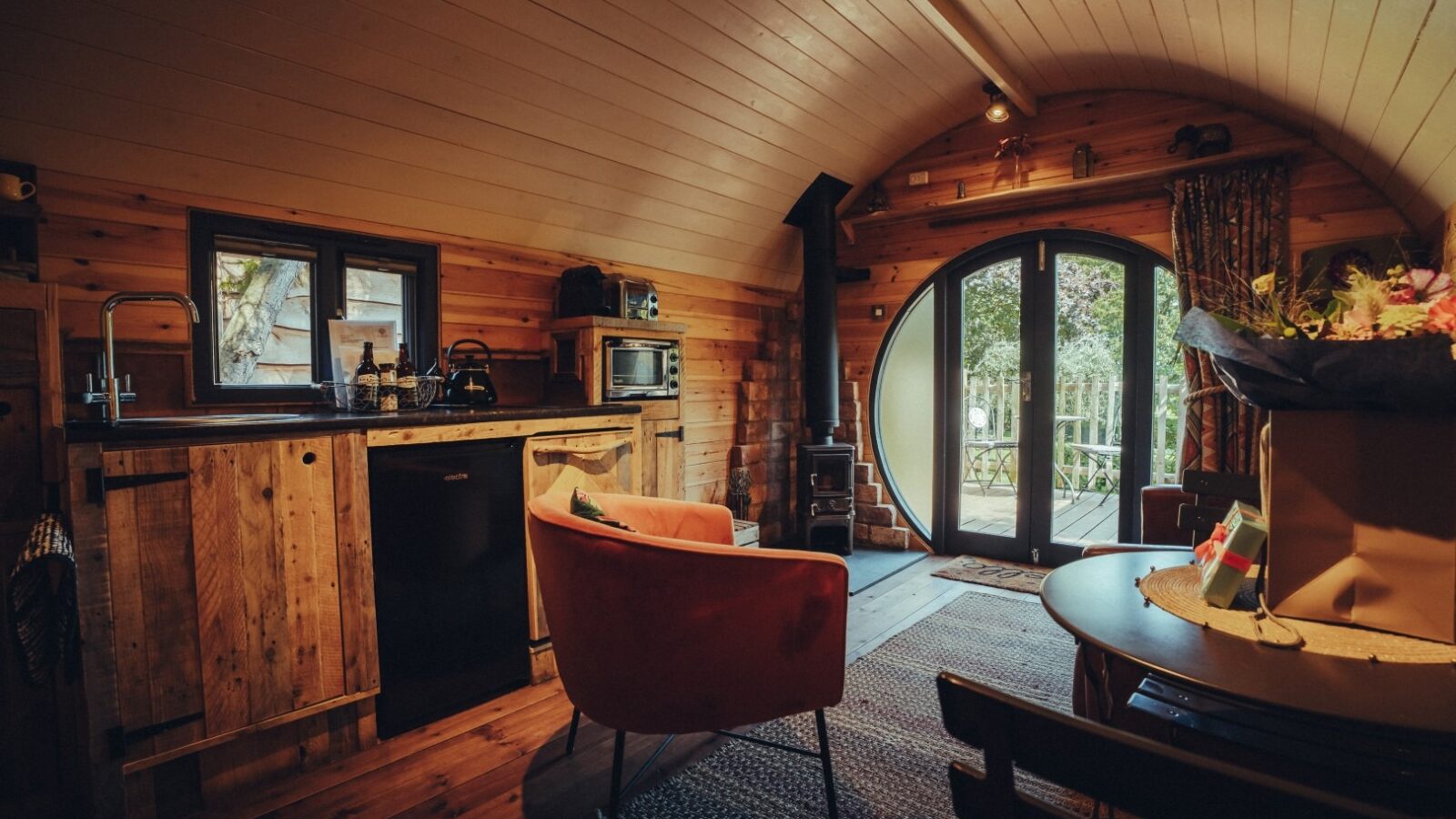 A cozy, rustic cabin interior with wooden walls and a curved ceiling. Features include a compact kitchen with a small refrigerator, sink, and microwave, a red armchair, a wooden table with chairs, and a circular glass door opening to a balcony overlooking the lush greenery of The Orchard.