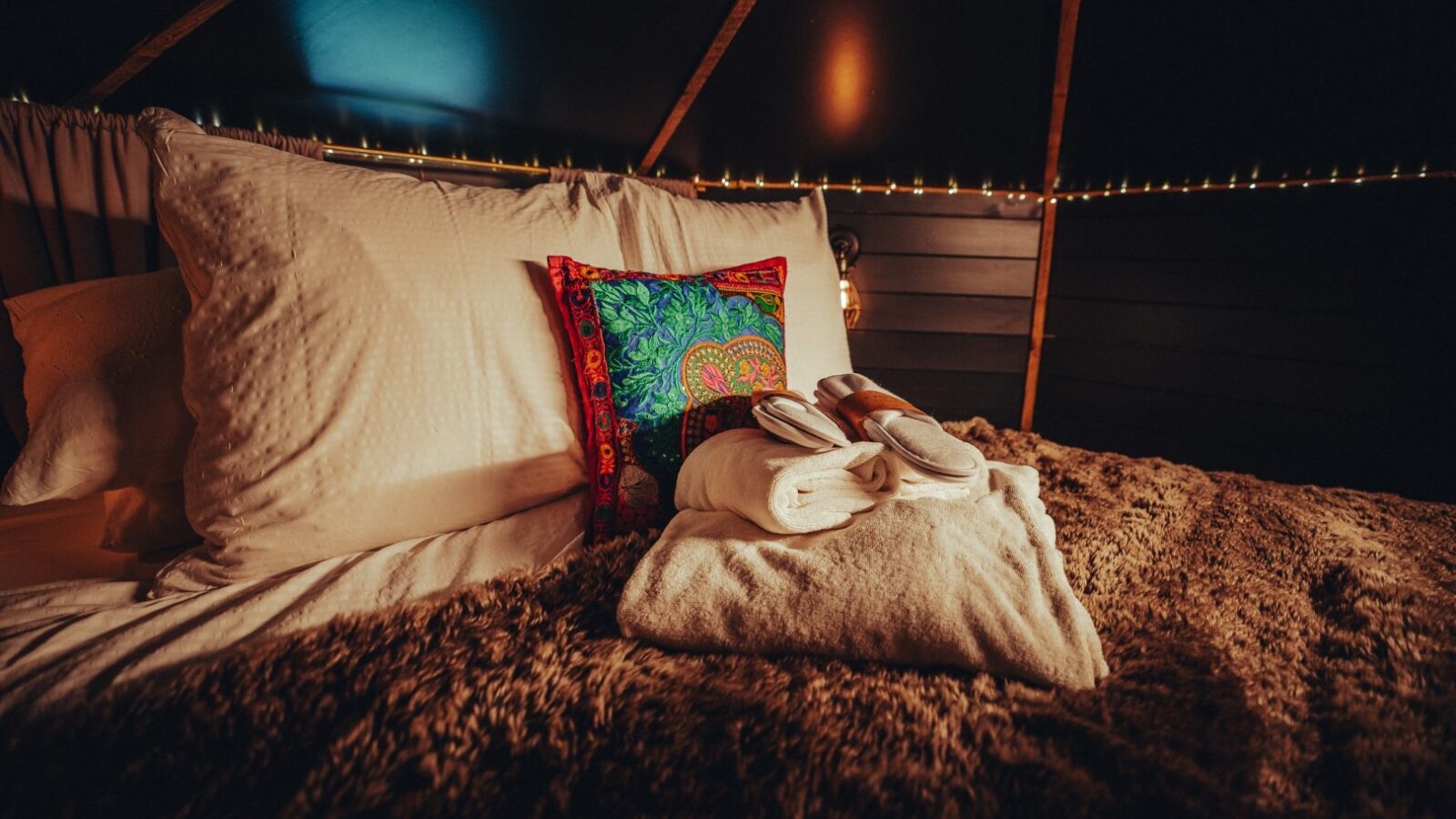 A cozy bed with white pillows and colorful decorative pillows under warm, dim lighting creates a welcoming space at Fenny Castle. Towels and slippers are neatly arranged on a fluffy, brown blanket. String lights adorn the dark walls, creating a tranquil and inviting ambiance reminiscent of The Orchard.