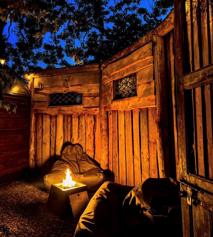 A cozy outdoor seating area at dusk with a wooden fence, two bean bag chairs, and a small fire pit. The warm glow from the fire illuminates the rustic wood surroundings of The Orchard while tree branches frame the darkening blue sky above.