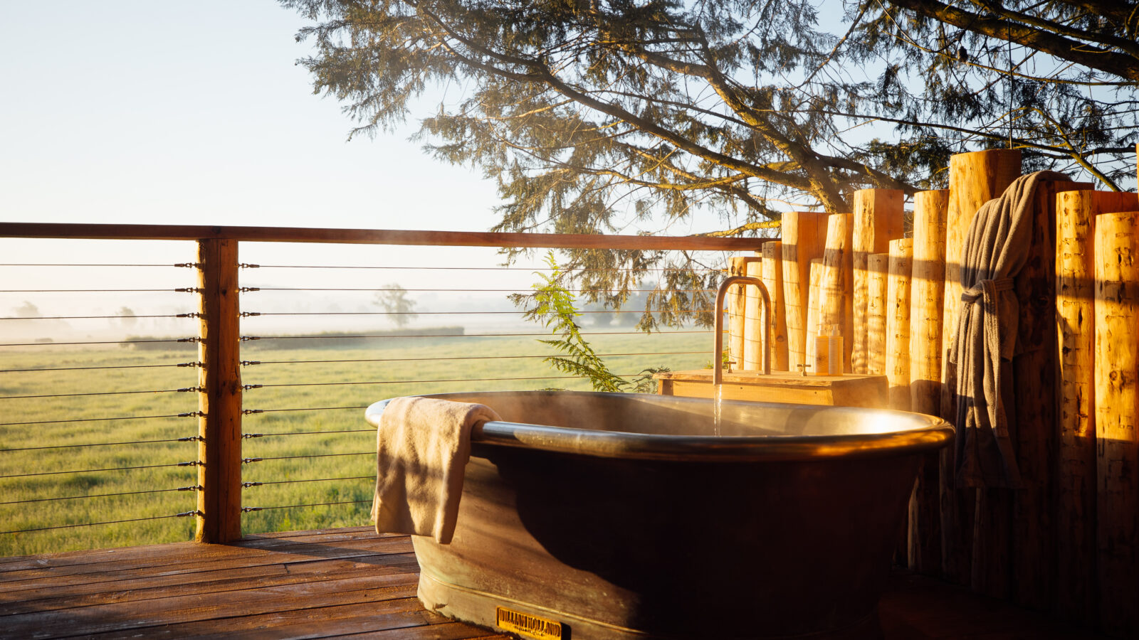 An outdoor copper bathtub on a wooden deck overlooks a scenic, foggy meadow at sunrise near The Orchard. A towel hangs over the tub's edge, and steam rises from the hot water. A rustic wooden fence and trees add to the serene, natural setting, hinting at views of Fenny Castle in the distance.