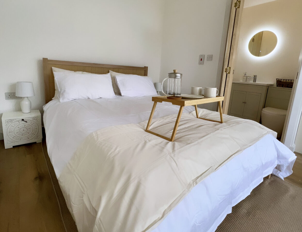 A cozy bedroom at The Perch features a neatly made bed with white linens and a wooden headboard. A breakfast tray with a carafe, two cups, and a bowl is placed on the bed. There is a white nightstand to the left and an open door leading to a bathroom on the right.