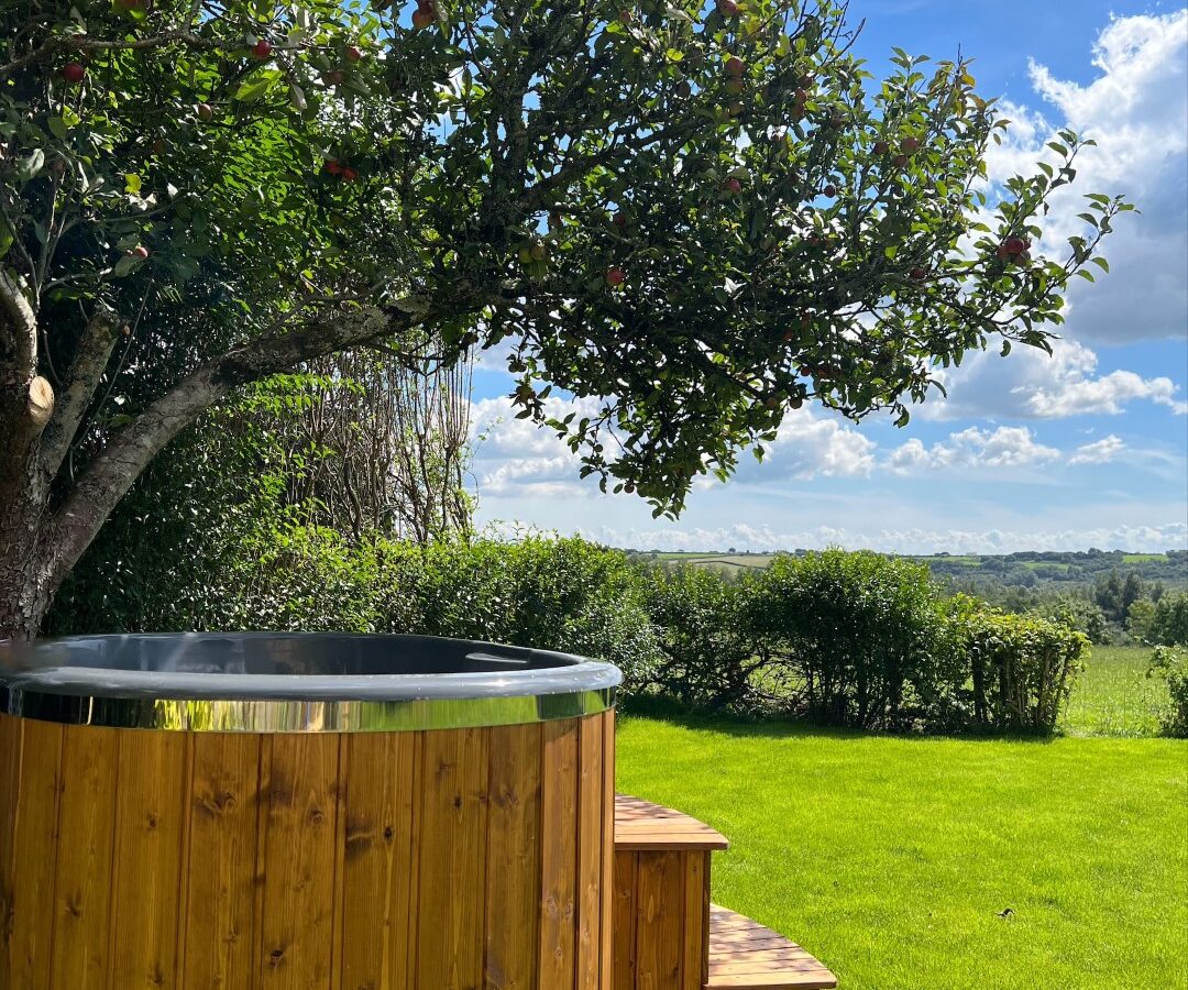 A wooden hot tub with steps, named The Perch, is placed under a tree in a lush green garden. The tree has apples hanging from its branches. A scenic view of rolling hills and a partly cloudy blue sky is visible in the background.