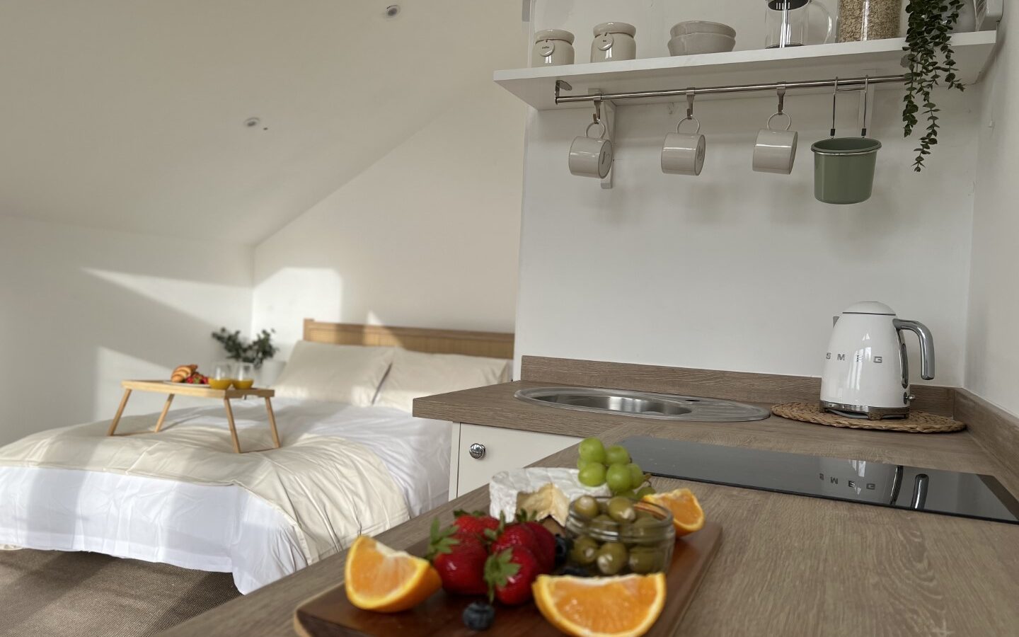 A modern and cozy bedroom at The Perch features a double bed with white bedding. In the foreground, a wooden tray on the kitchen counter holds assorted fruits like oranges, grapes, strawberries, and blueberries, along with some olives. Shelves and kitchen items are visible.