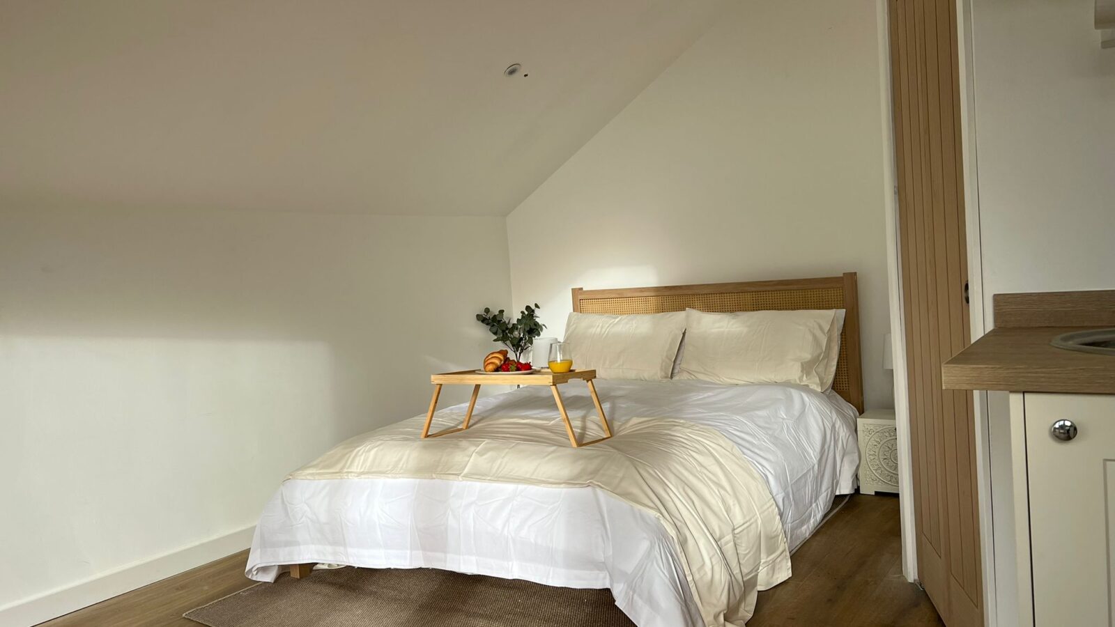 A cozy bedroom at The Perch features white walls and a double bed adorned with a white duvet and pillows. A wooden tray holding an orange, a cup of tea, and a small plant rests on the bed. The room boasts a wooden floor and a small kitchenette visible on the right side.