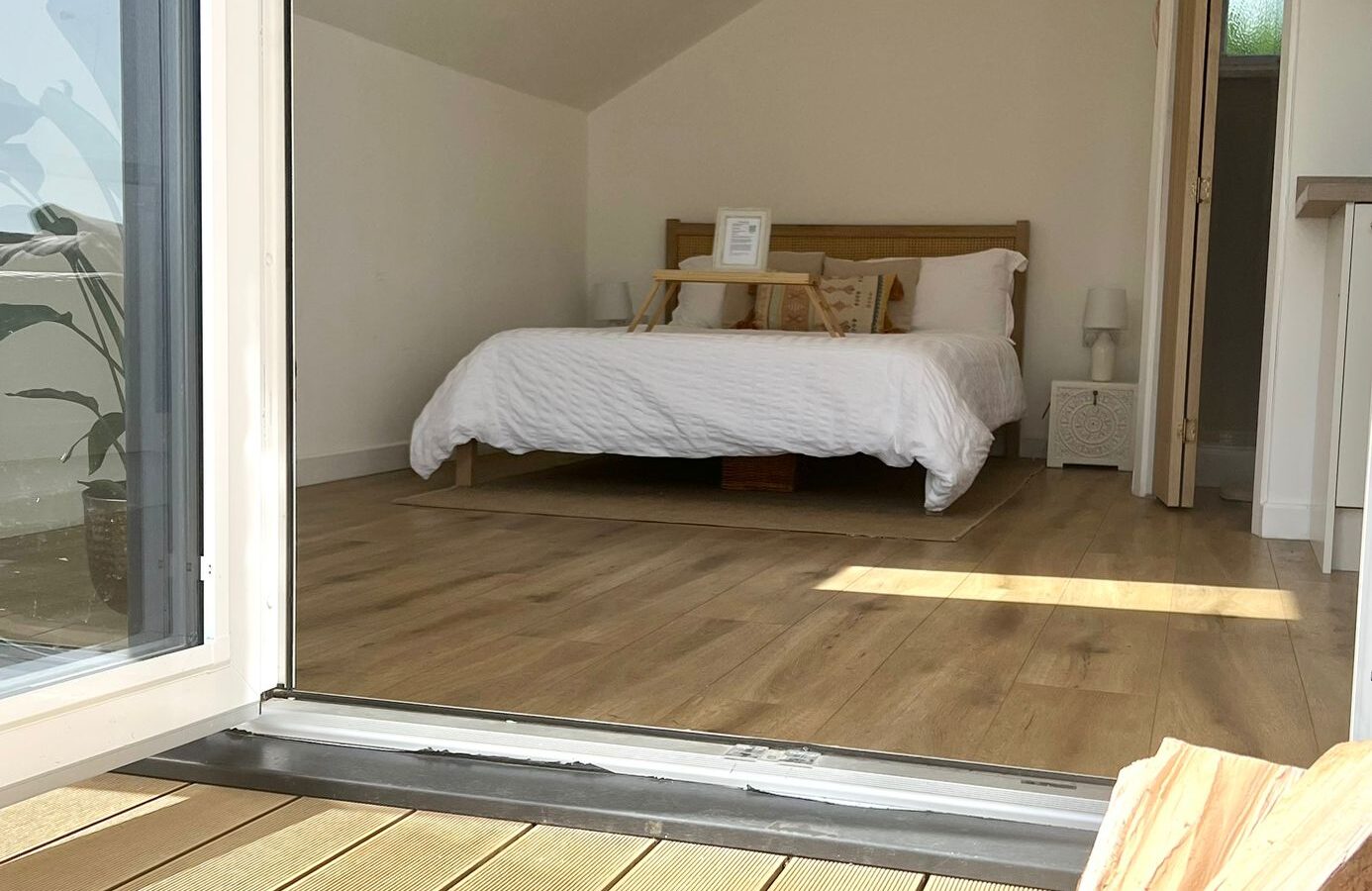 A cozy bedroom with a sloped ceiling and recessed lights, featuring a neatly made bed with white linens and wooden headboard. Wooden flooring extends to a small balcony called The Perch, where a basket of firewood is visible in the foreground. A door is slightly open, inviting natural light.