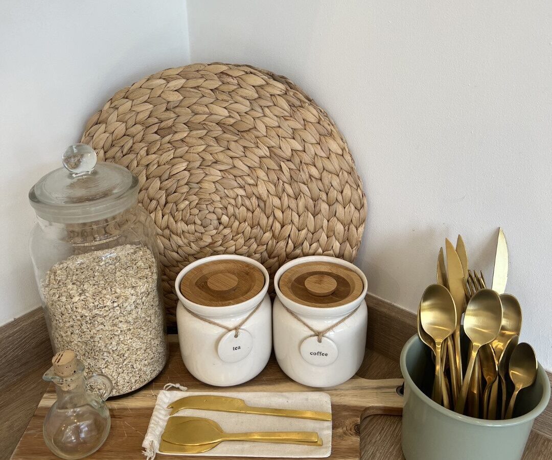 A kitchen counter at The Perch features a glass container of oats, two labeled ceramic jars (one for tea, another for coffee), golden cutlery in a green container, a small textured jar with a cork lid, and a woven placemat leaning against the wall.