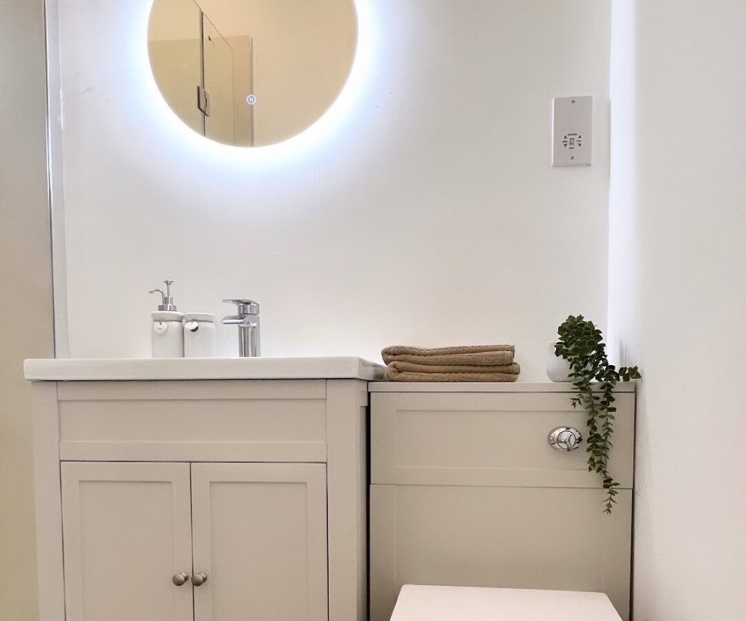 A modern bathroom, known as The Perch, features a white vanity with a round illuminated mirror above it. There is a small white sink with a silver faucet, a folded towel, and a plant on the countertop. Adjacent to the vanity is a sleek, white toilet. The floor is wooden.