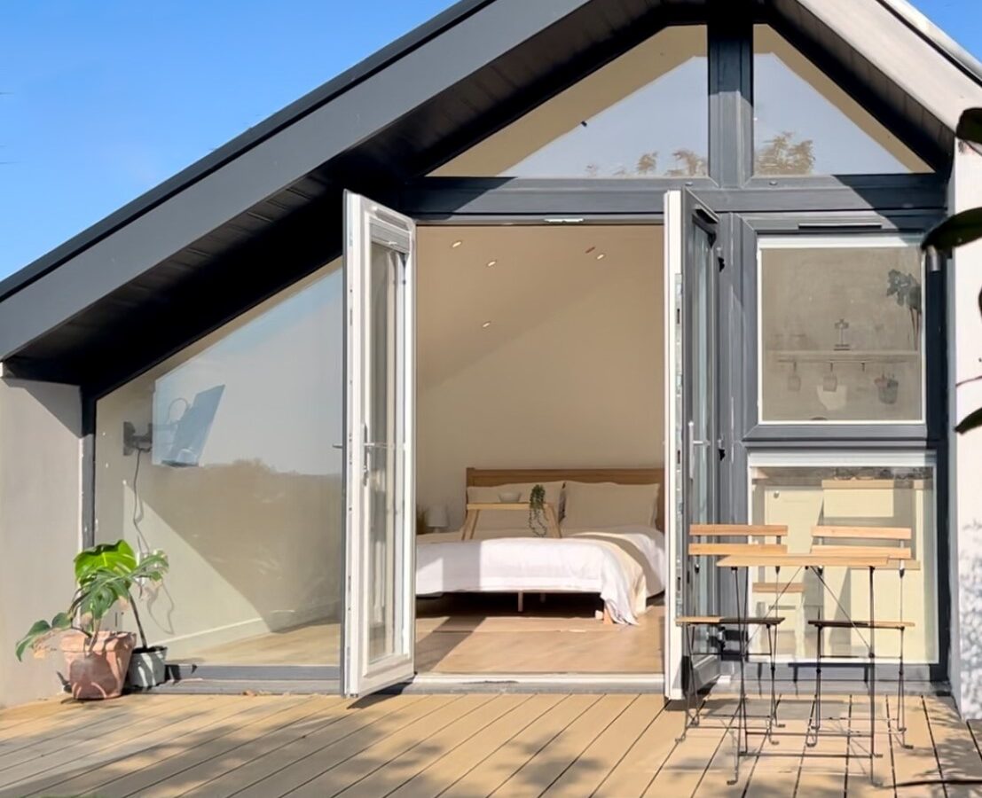 A cozy small house features a wooden deck with a potted plant and a bistro set, perfect for dining al fresco. The house has large, open glass doors revealing a bright interior with a neatly made bed and minimal decor. The roof is sloped, and the scene is set against a clear blue sky.