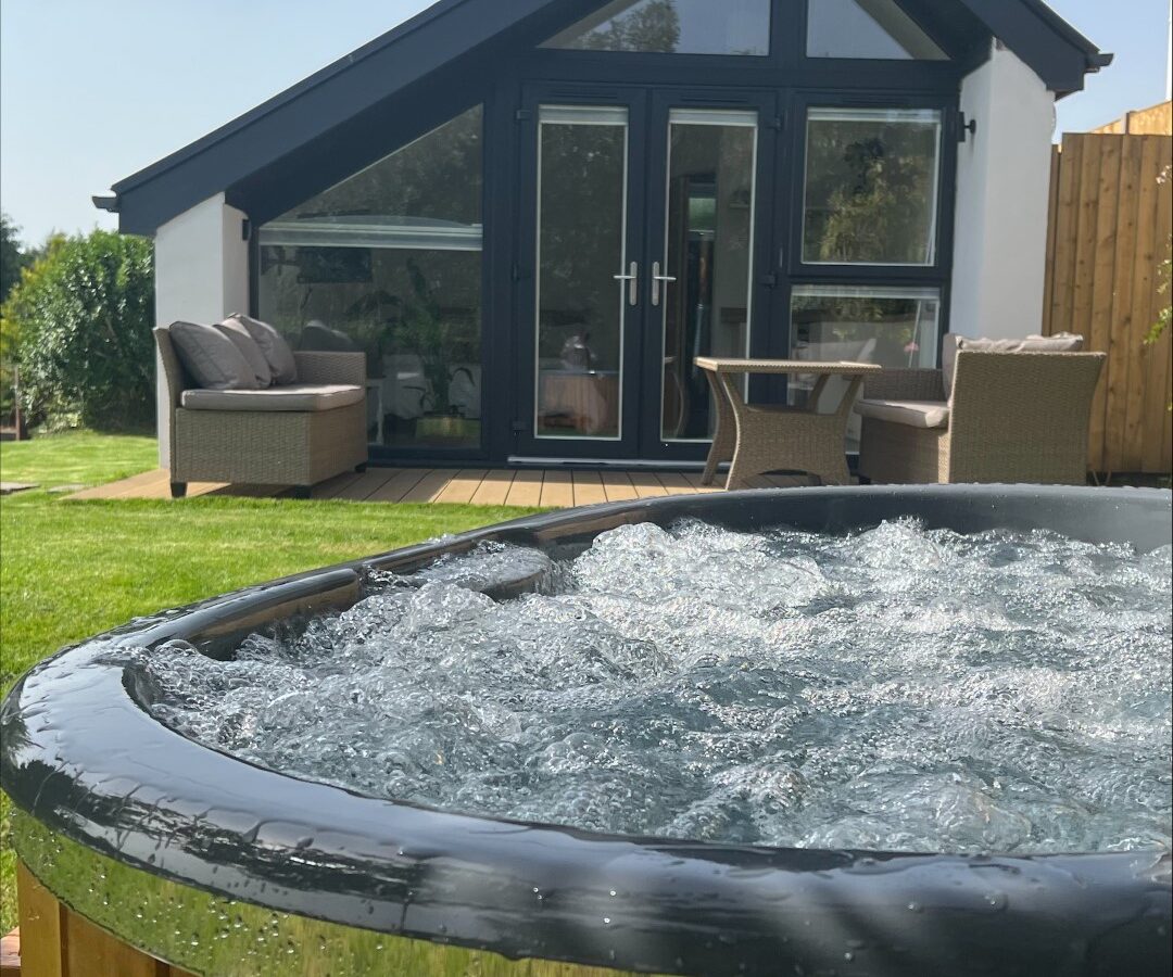 A wooden hot tub with bubbling water sits in the foreground. Behind it, The Perch, a cozy cottage with large glass doors and windows, features an outdoor patio with wicker furniture and cushions. The setting is under a clear blue sky with green grass and some trees visible.