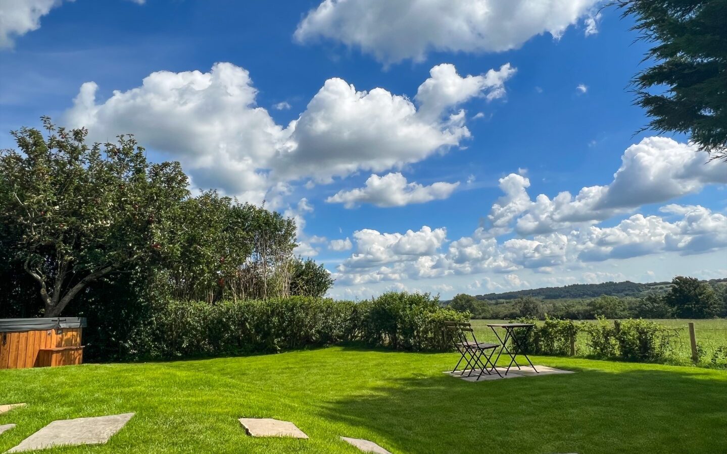 A lush green yard under a blue sky scattered with fluffy clouds. On the right, The Perch features a set of purple outdoor furniture consisting of a table and two chairs. Trees and bushes line the yard's boundary, and stepping stones lead across the grass.