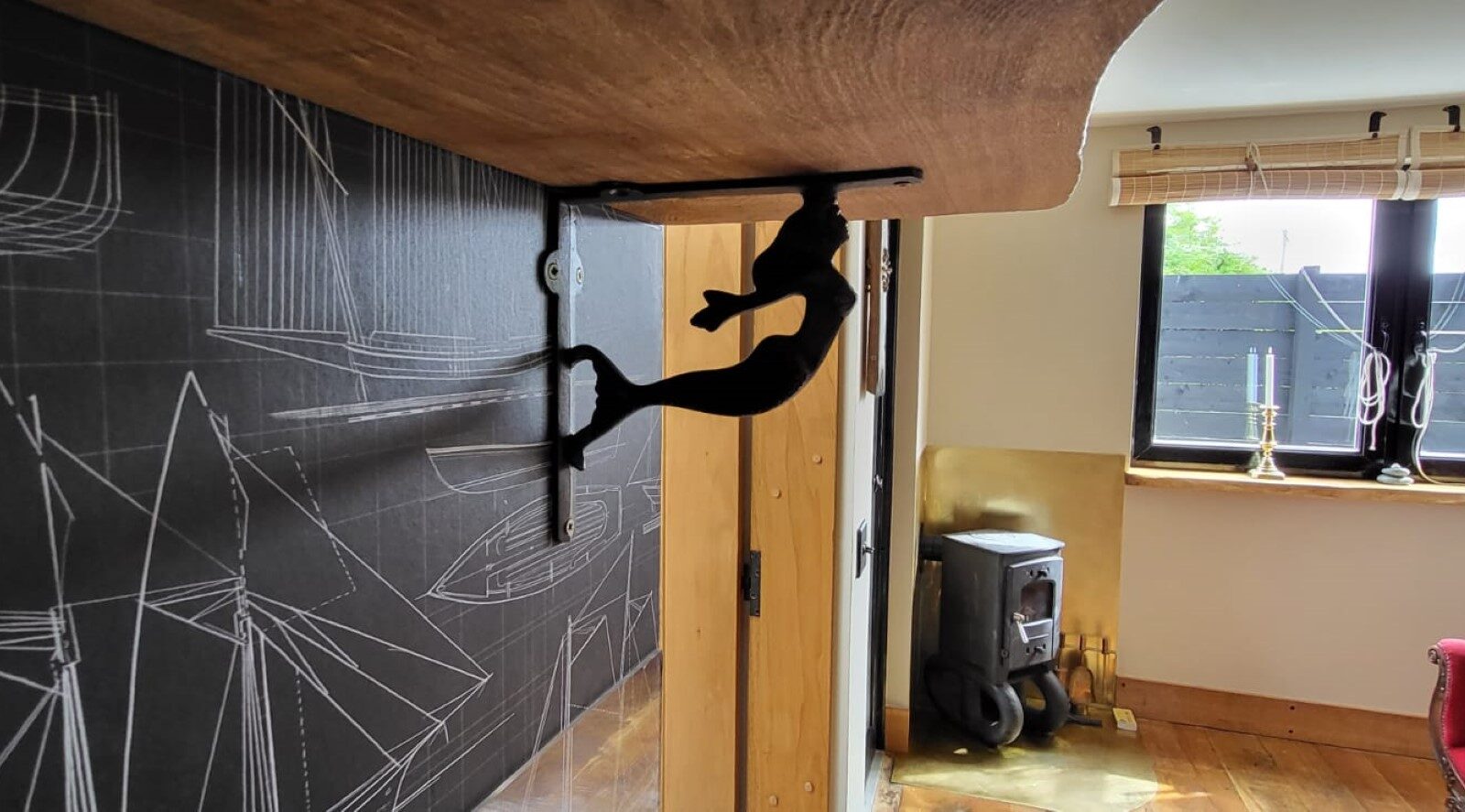 A wooden countertop supported by a decorative metal bracket in the shape of a rock climber. The side of the wall features a detailed blueprint-style wallpaper with sailboat designs, reminiscent of The Quarterdeck. In the background, there are windows with wooden blinds, a small wood-burning stove, and two candle holders.