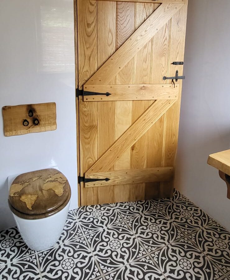 A rustic wooden door with black hinges and latch, atop a black and white patterned tile floor. To the left is a small wooden shelf with two hooks, and below it sits a round stool with a weathered seat. The room, known as The Quarterdeck, boasts a starry sky ceiling design.